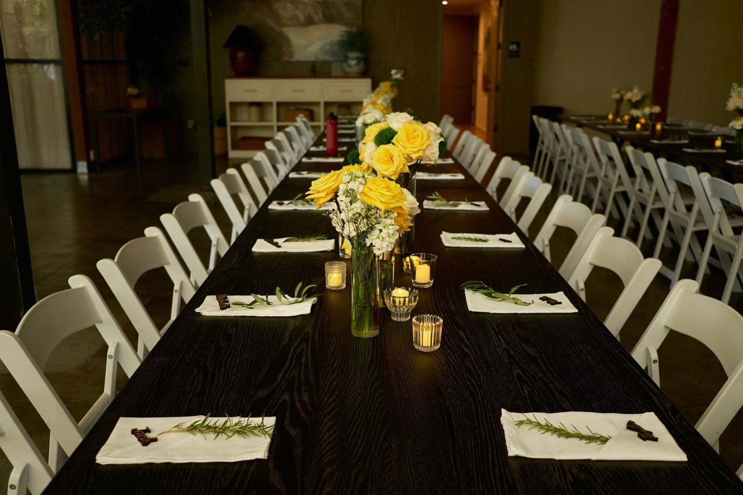 Long wedding reception table set with white folding chairs, yellow flowers, tea light candles, and napkins topped with greenery.
