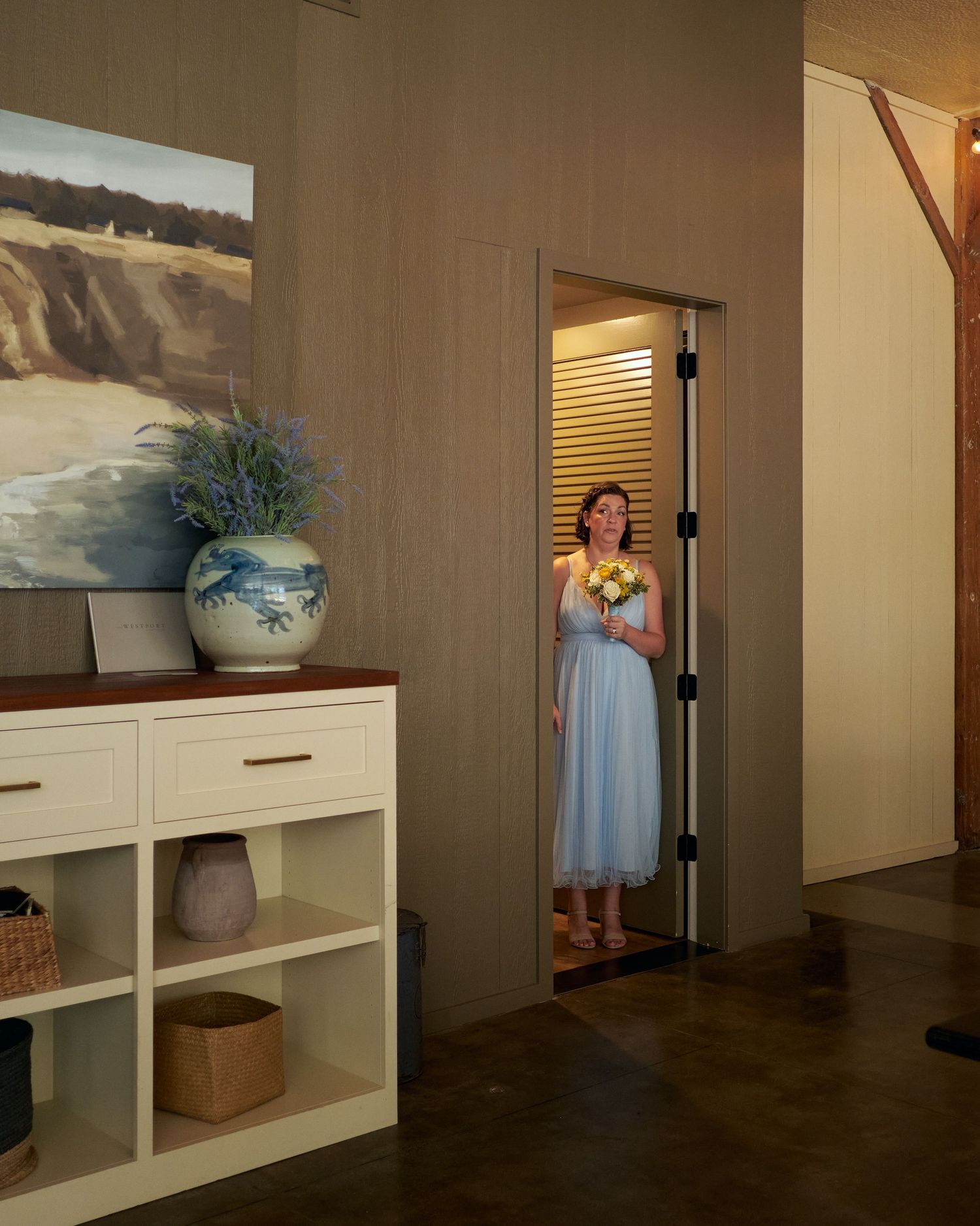 A bridesmaid in a light blue dress holding a small bouquet stands in a doorway near a coastal painting and vase.