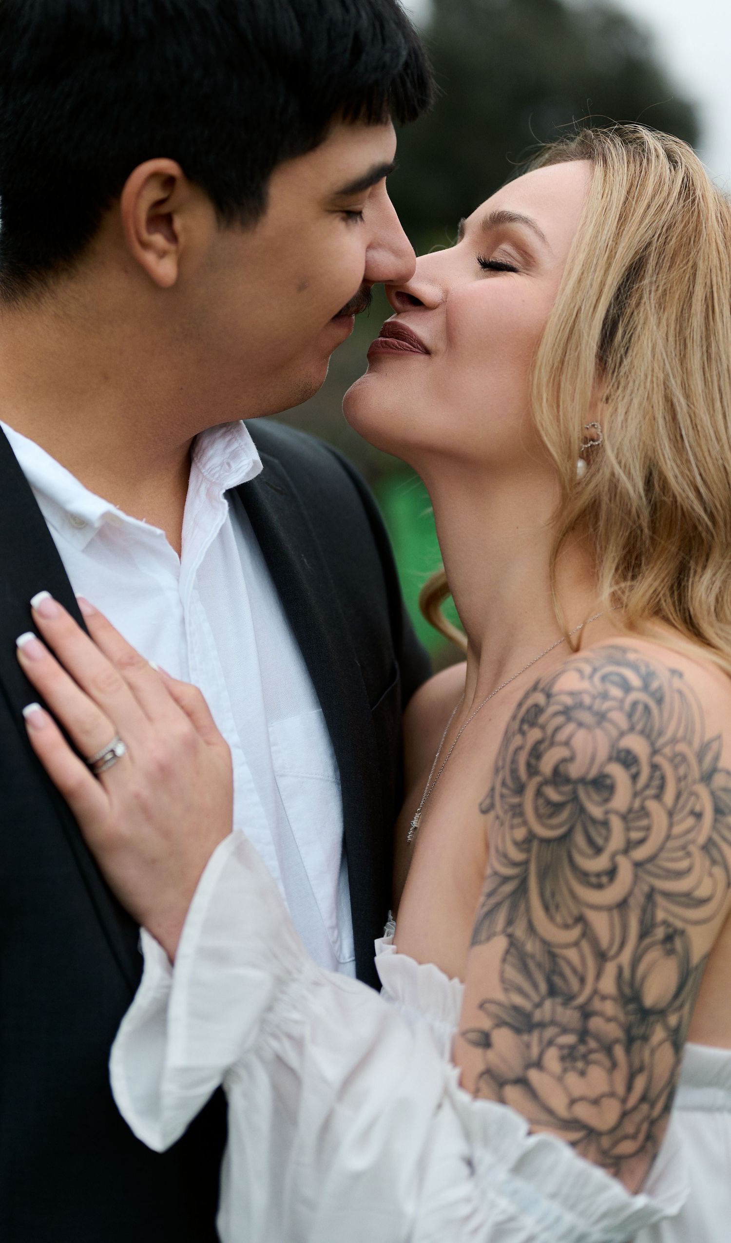Engaged couple shares an almost-kiss as the woman, wearing a ring and a floral shoulder tattoo, rests her hand on his chest.