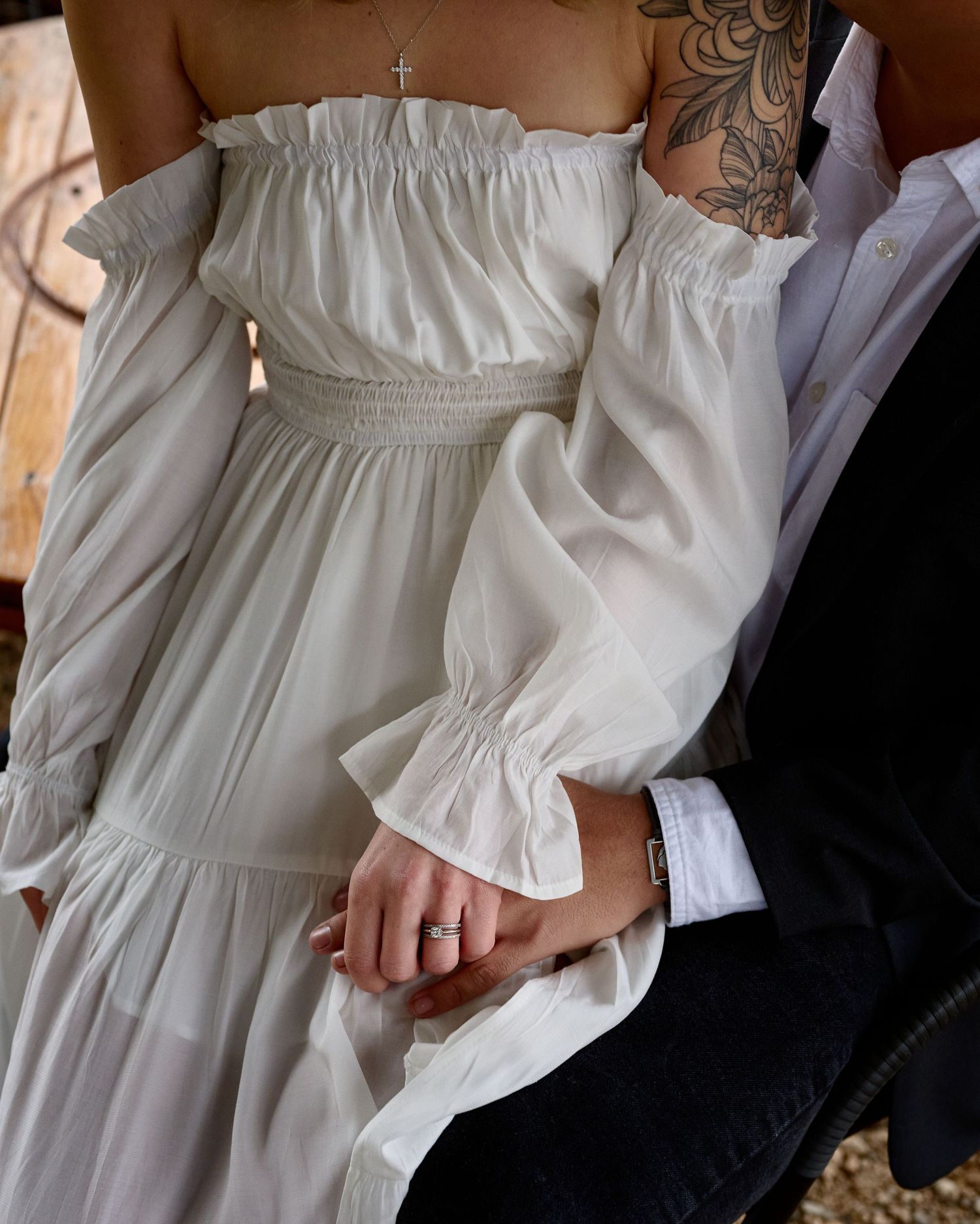 Close-up of a bride in a white off-shoulder dress with a floral tattoo holding hands with her groom, her ring visible.