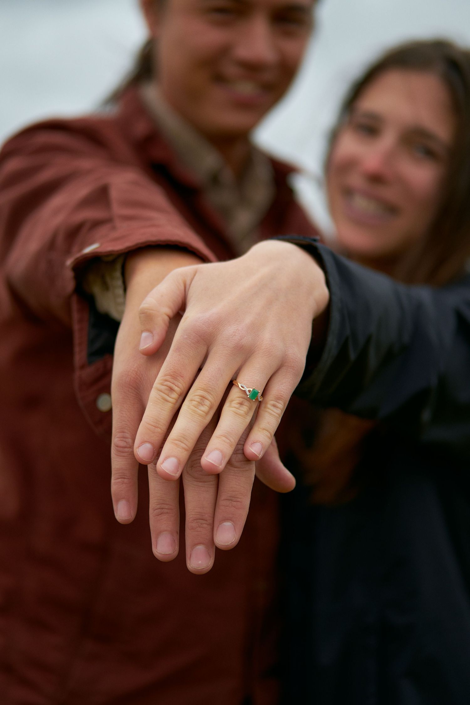 A smiling couple stands outdoors with blurred faces as the foreground focuses on a hand wearing a green gemstone engagement ring.