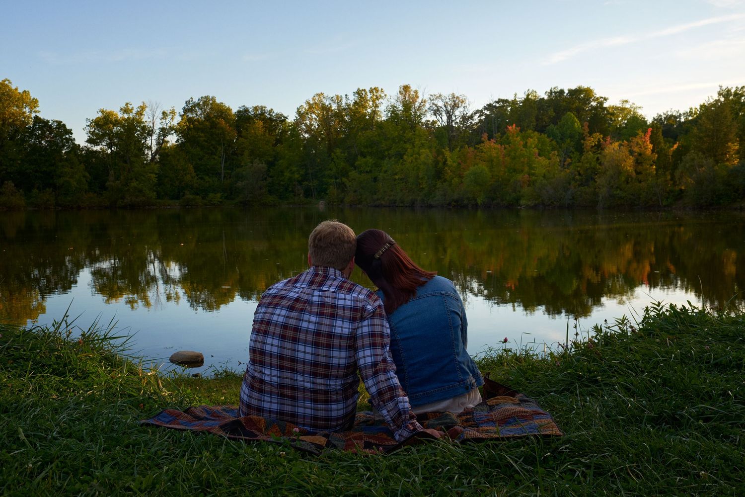 A couple sits close together on a picnic blanket by a calm lake, watching sunset light reflect on the water and fall trees.