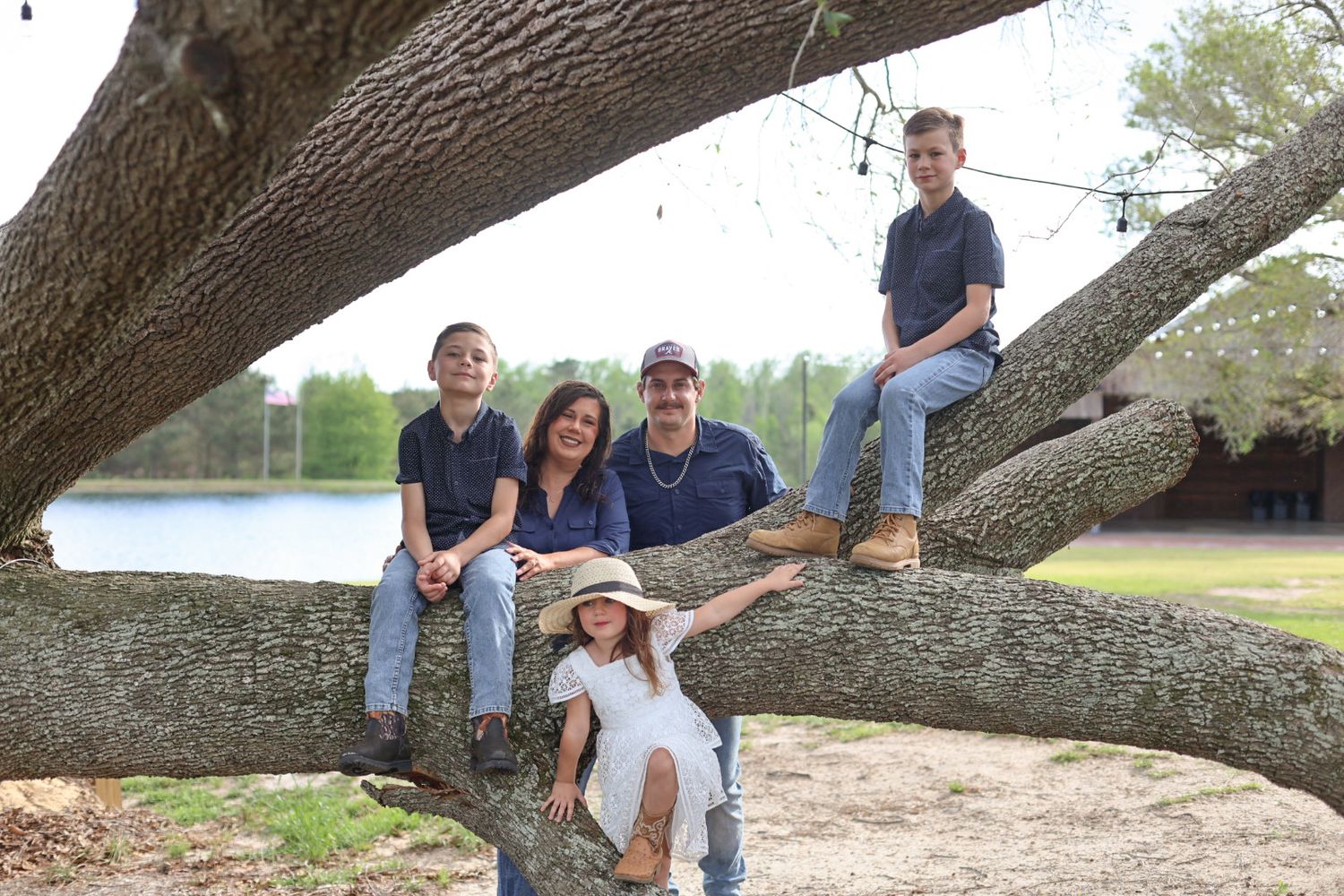 Family of five posing on the large branches of a mossy oak tree beside a lake, with a young girl in a sun hat.