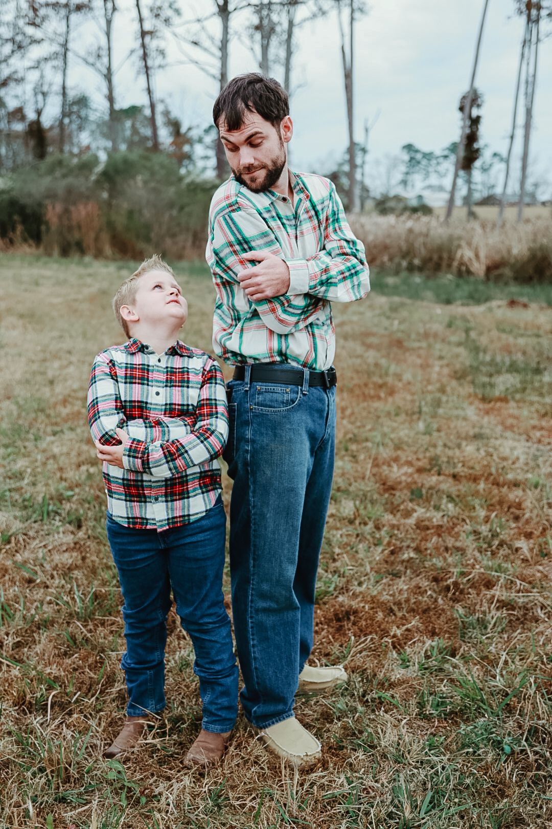 A bearded man and a young boy in matching plaid shirts stand with crossed arms in a grassy field, the boy looking up at him.