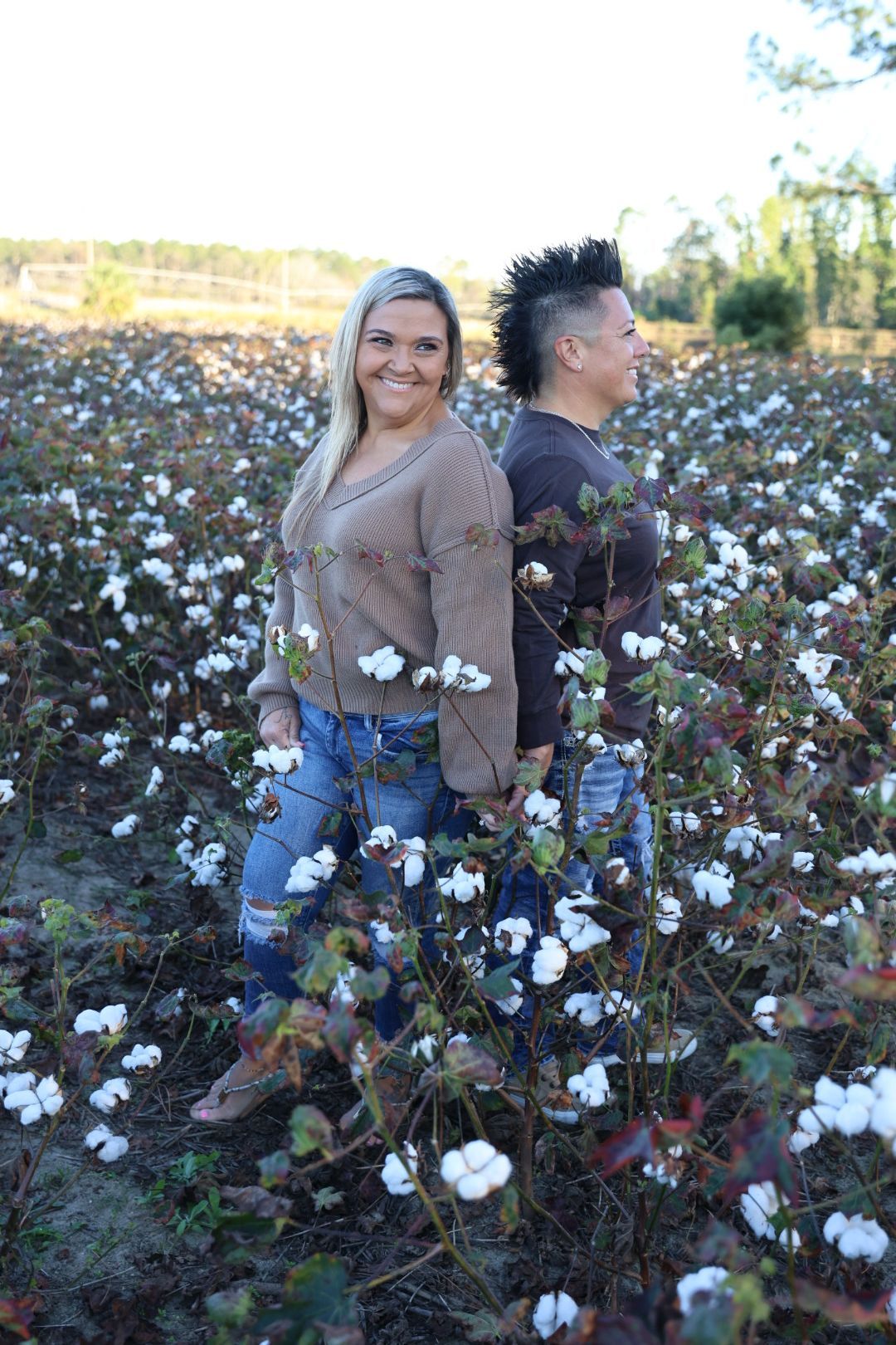 Two smiling women in sweaters and jeans stand back to back in a sunlit cotton field with white cotton bolls in bloom.