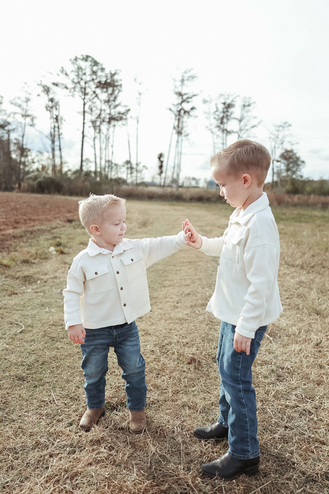 Two young boys in cream shirts and jeans hold hands while facing each other in a golden field with trees behind them.