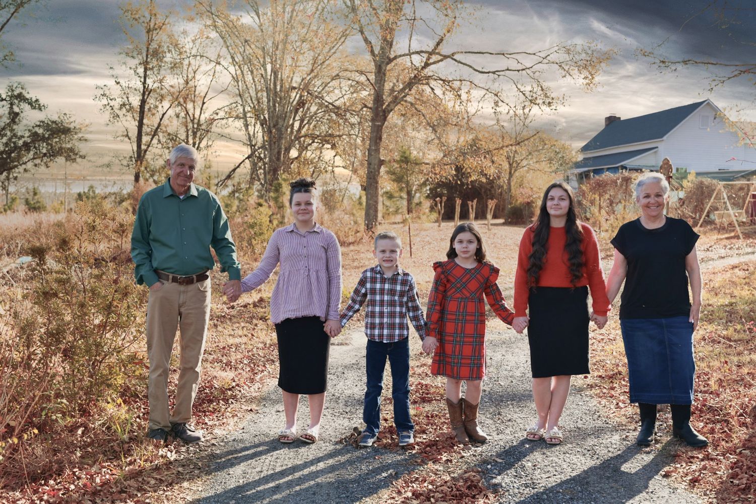 Multigenerational family of six holds hands while standing on a gravel path lined with autumn trees near a country home.