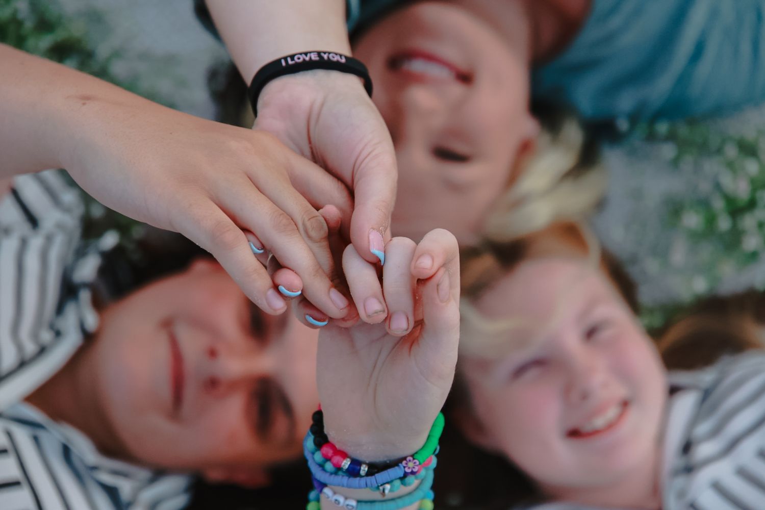Overhead view of hands stacked together in the center, with three smiling people lying on the grass and colorful bracelets visible.