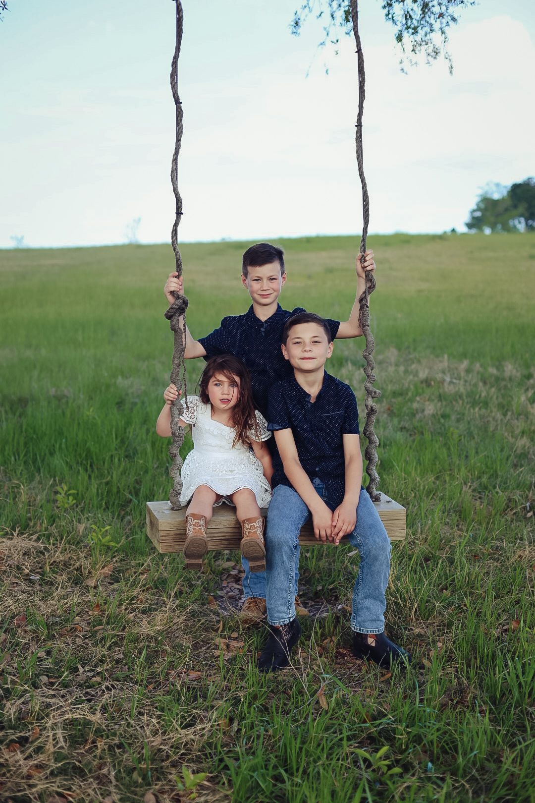 Three children sitting and standing together on a wooden rope swing in an open green field under a soft sky.