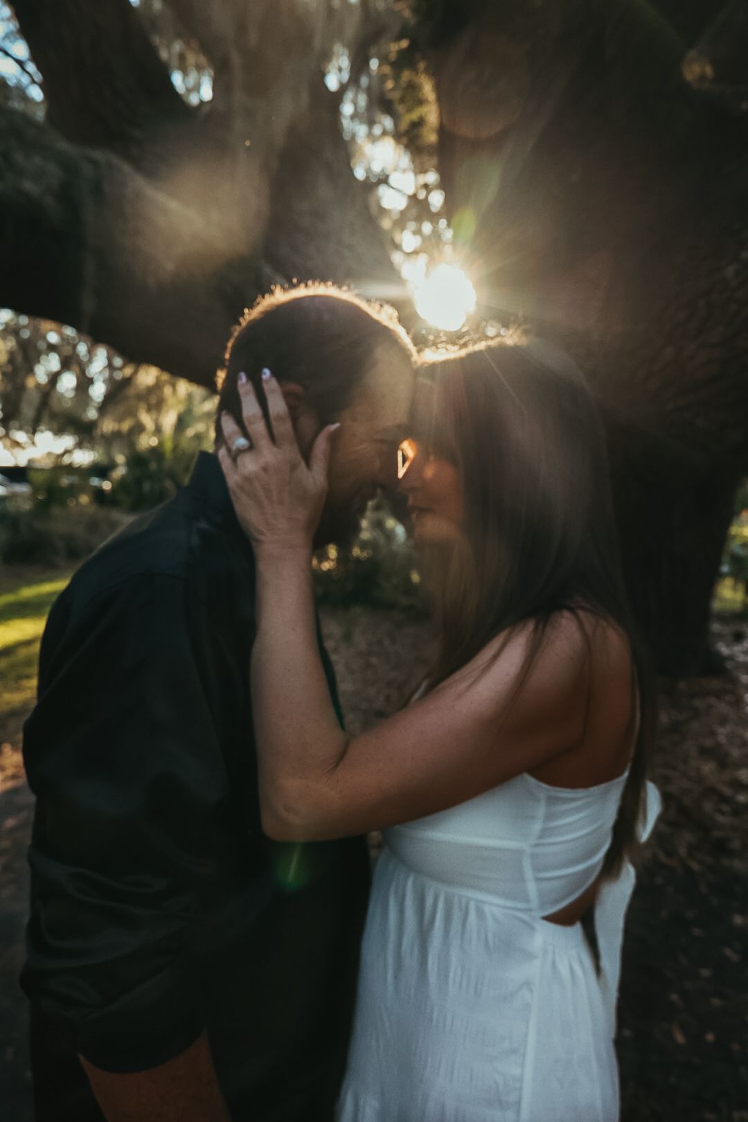 A couple shares a romantic moment beneath a large oak tree as golden sunlight flares through the branches behind them.