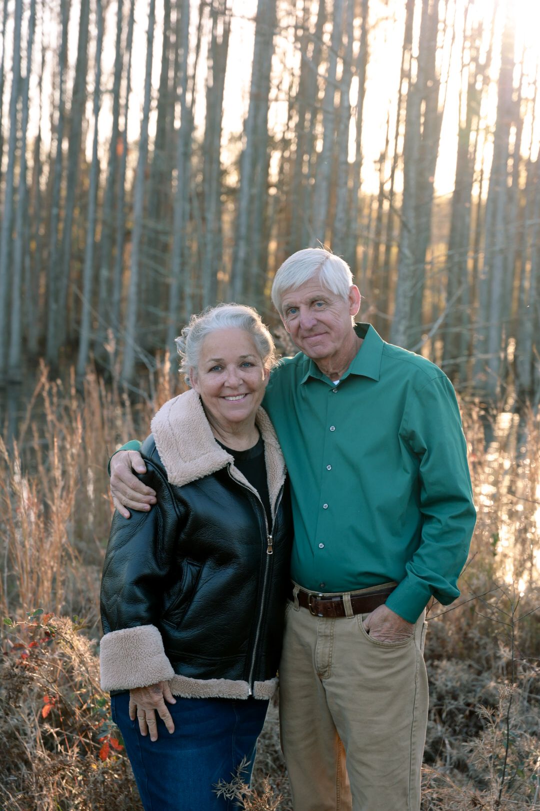 Smiling older couple in a green shirt and shearling-trimmed leather jacket pose arm in arm in a sunlit wooded field.