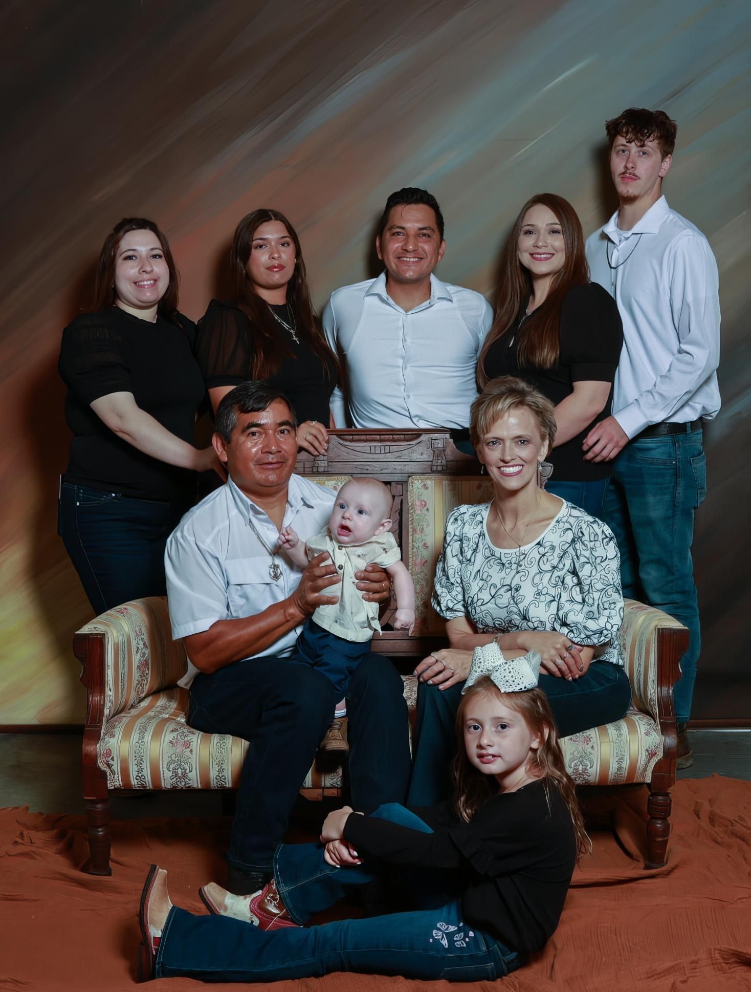 Multigenerational family portrait with parents, grandparents, adult children, a baby and a young girl posed around a vintage sofa.