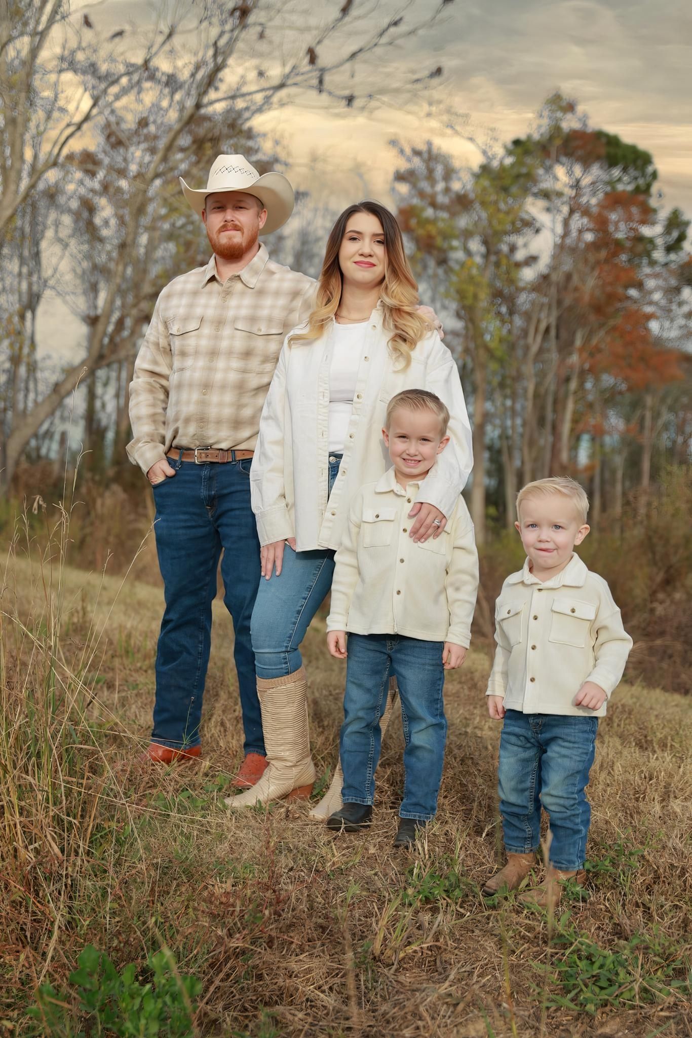 A family of four in denim and cream outfits poses in an autumn field, with the father wearing a cowboy hat and two young boys.