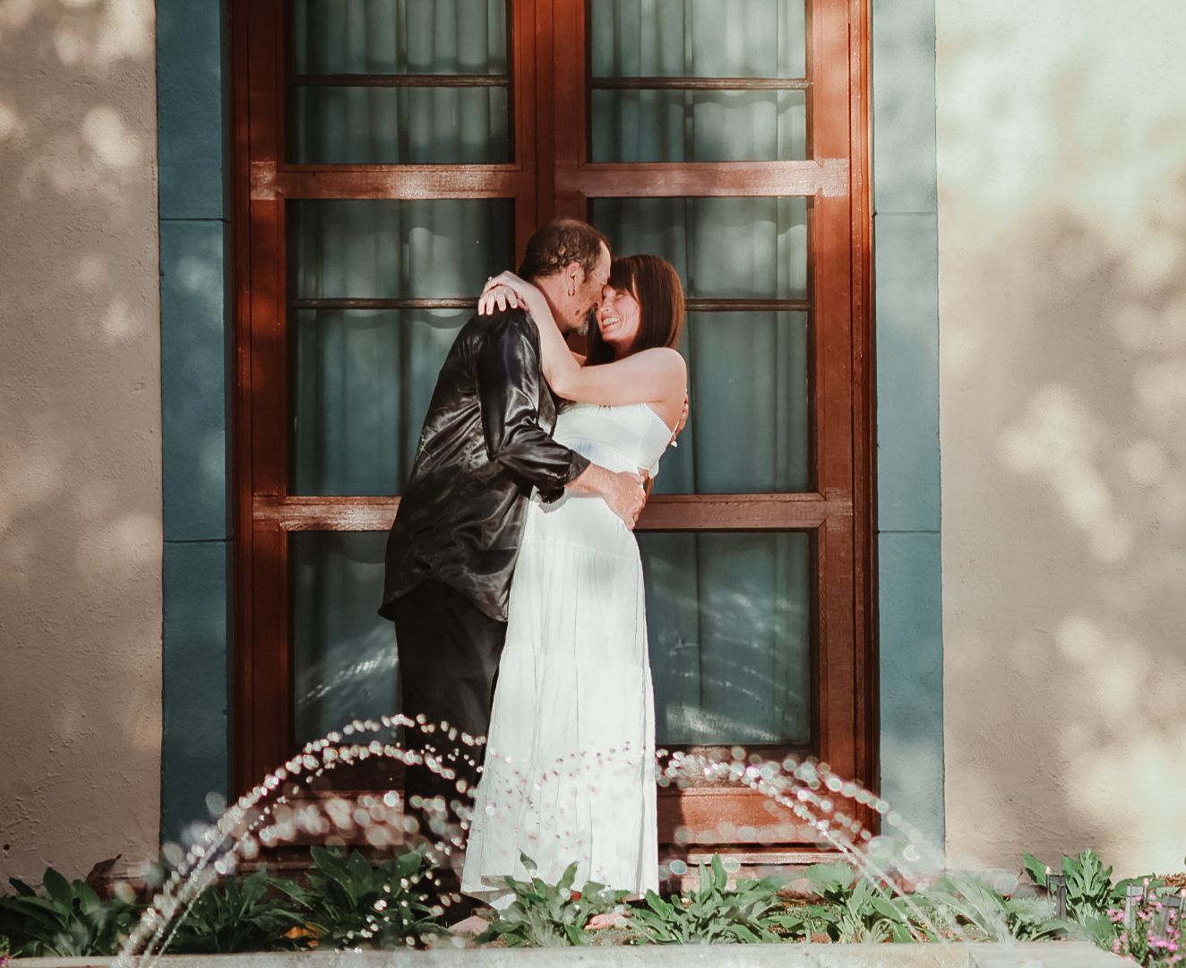 Couple embrace and kiss in front of a tall wooden window with a fountain spraying in the foreground.