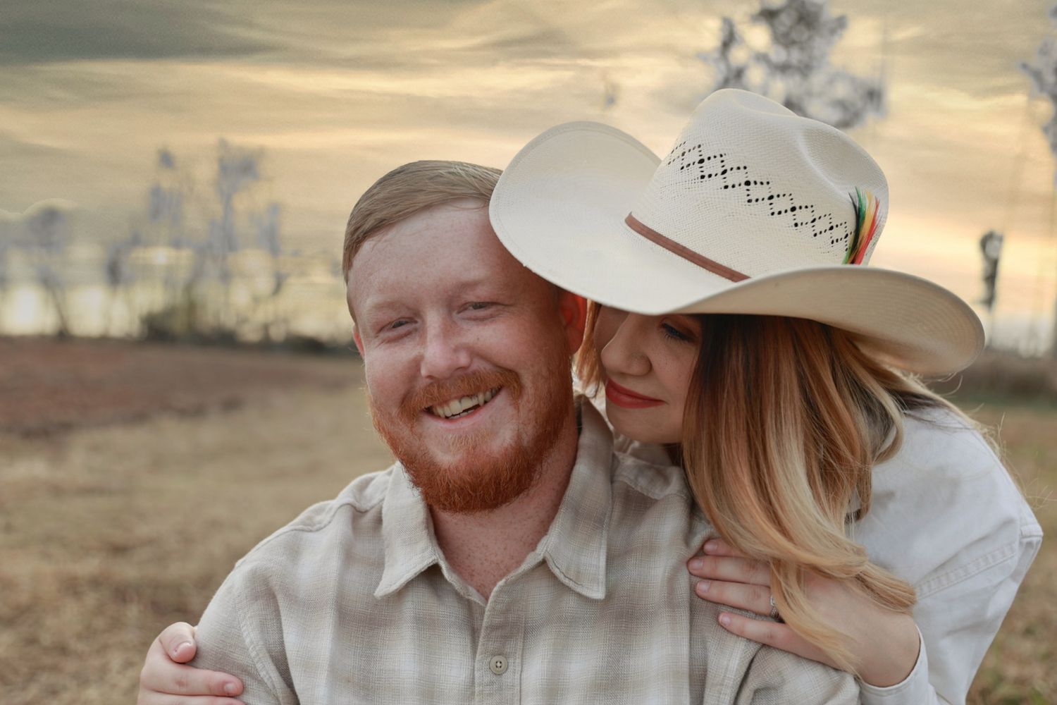 Smiling bearded man hugs his partner, who wears a cowboy hat, against a golden sunset sky in an open field.