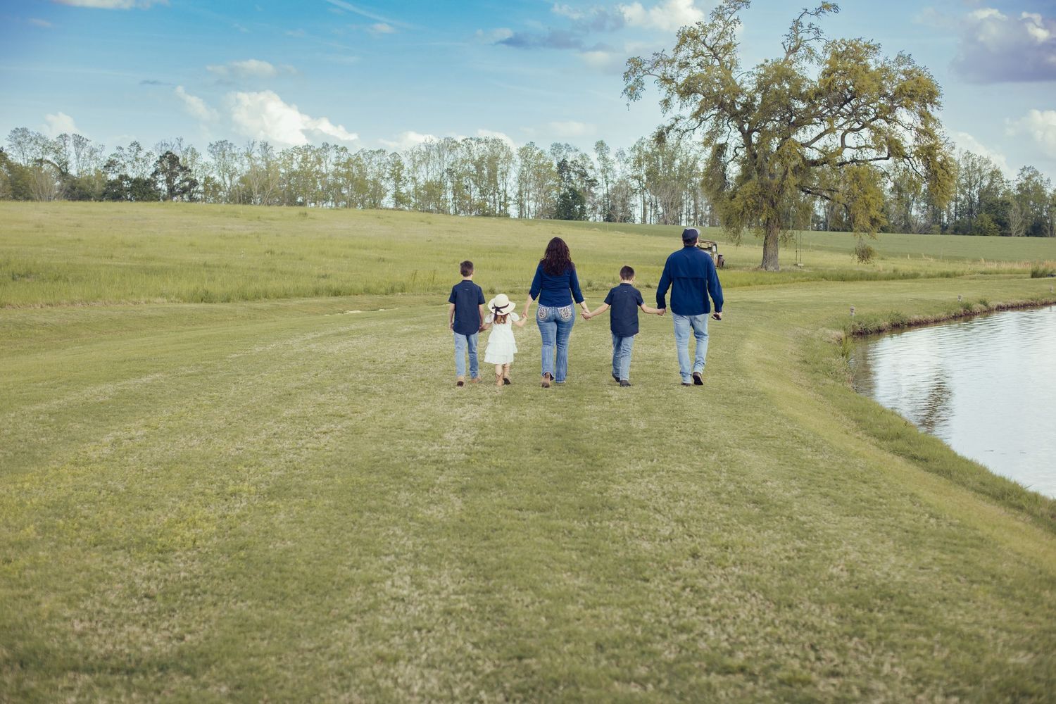 Family of five walking away on a grassy lakeside path toward a tree, holding hands on a bright sunny day.