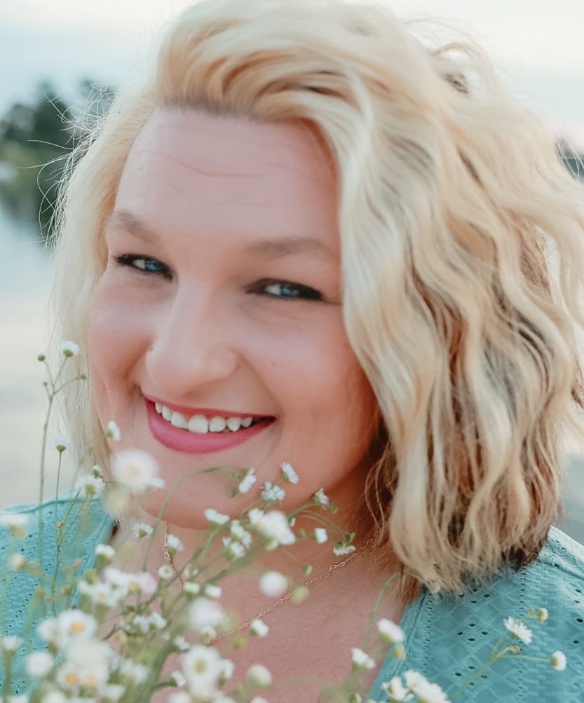 Smiling woman with wavy blonde hair holding delicate white wildflowers beside a calm lake at sunset.