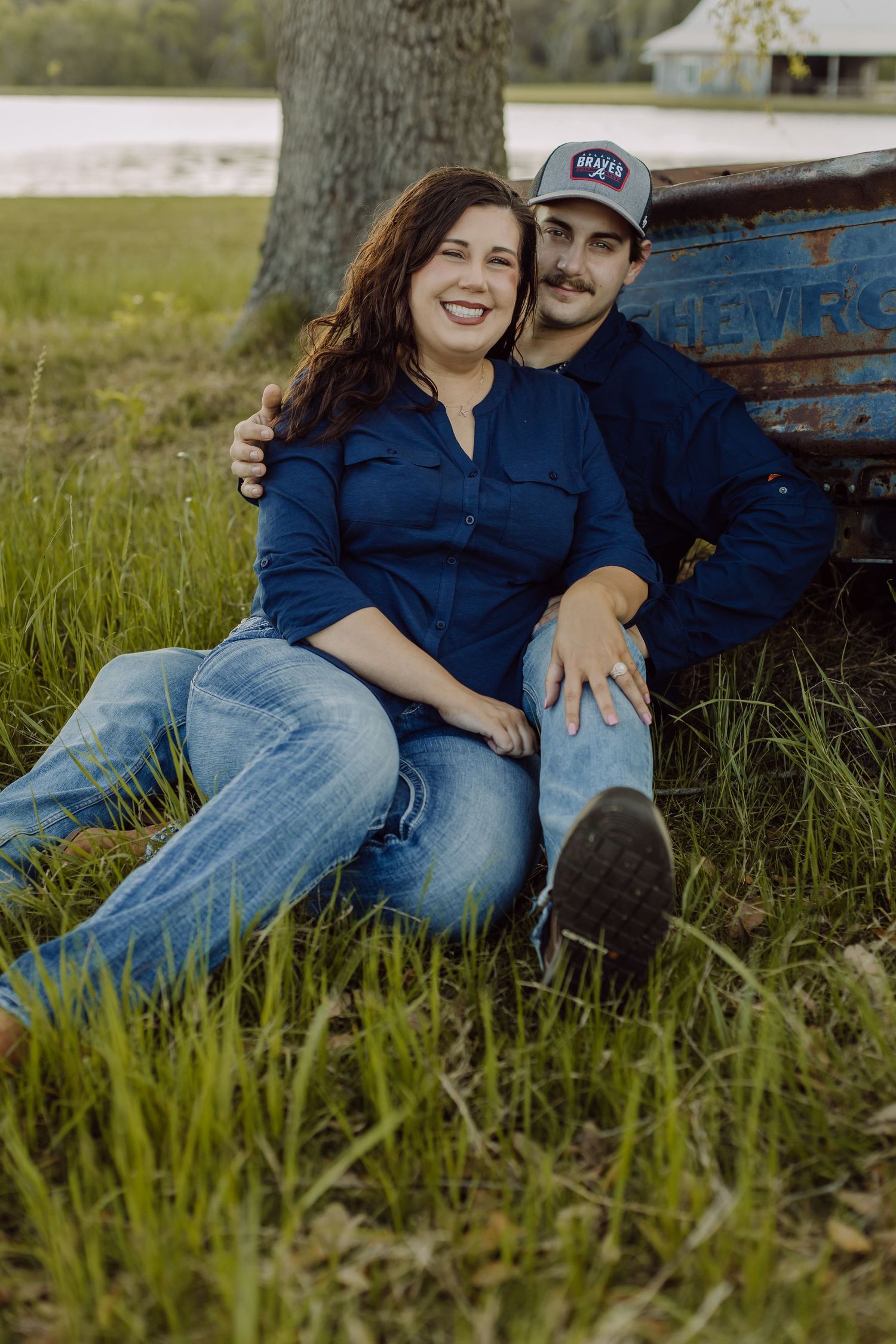 Smiling woman and man in a ball cap sitting together in the grass beside a vintage blue truck near a lake.