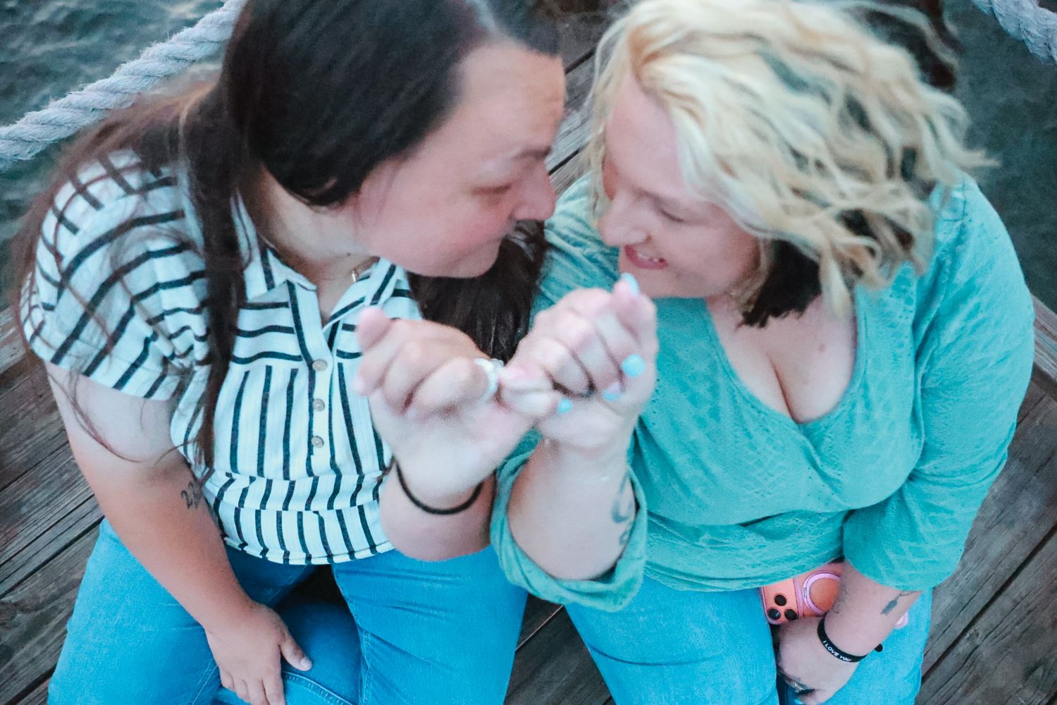 Two women in matching blue tones smile at each other while holding hands on a wooden dock by the water.