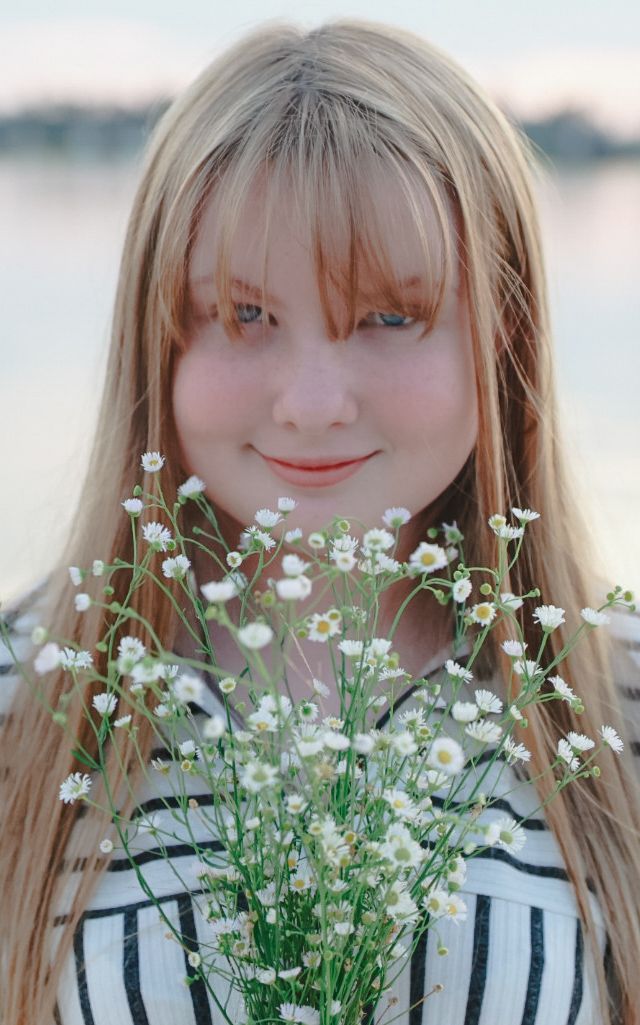 A young woman with long blonde hair smiles softly while holding a bouquet of small white wildflowers by the water.