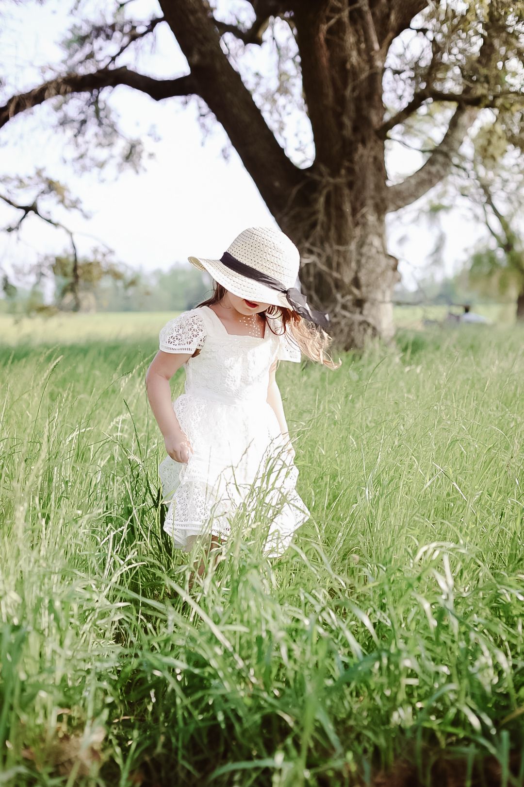 Young girl in a white lace dress and straw sun hat walking through tall green grass near a large oak tree.