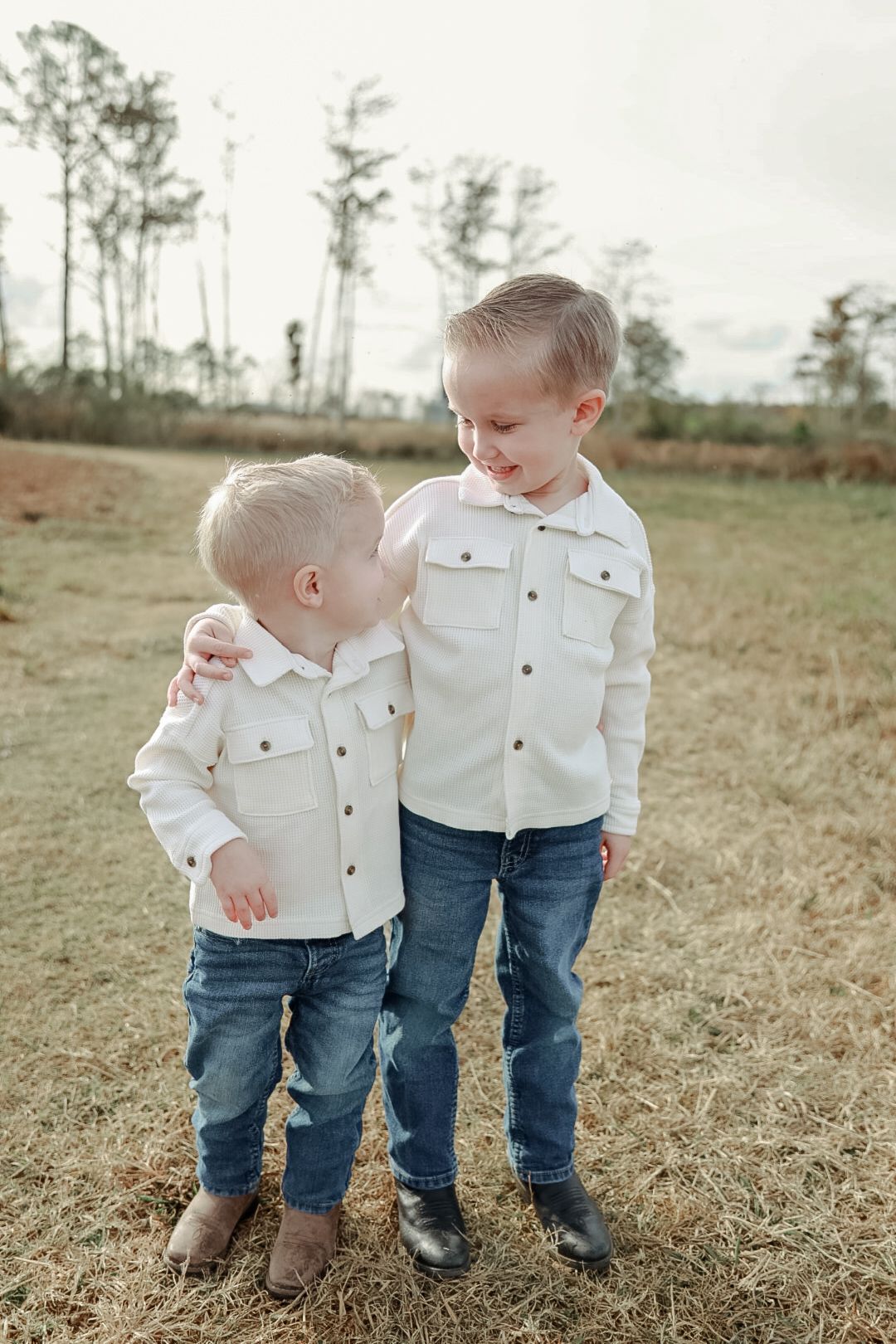 Two little brothers in cream button shirts and blue jeans hug each other while standing in an open field with tall trees.
