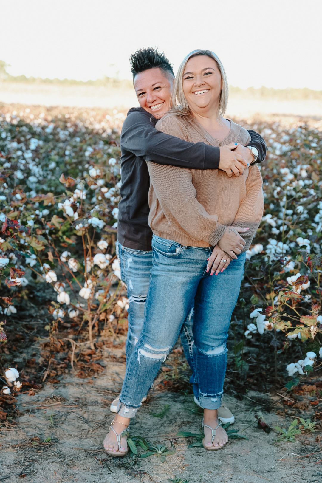 Two smiling women in jeans and sweaters hug from behind while standing in a blooming cotton field at golden hour.