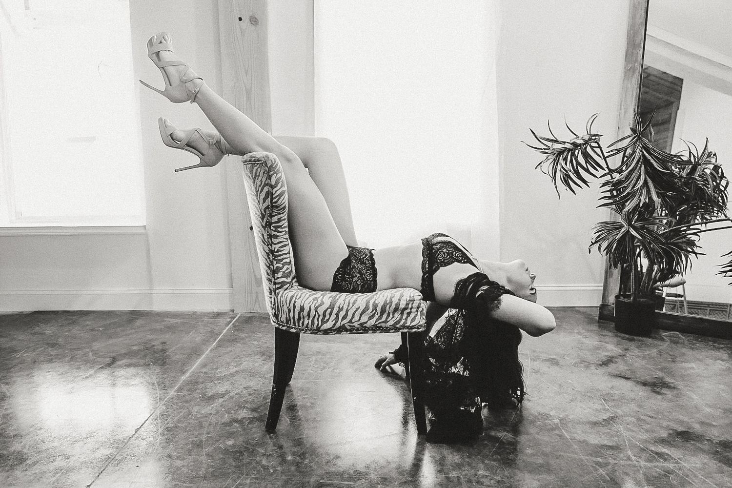Black and white boudoir photo of a woman reclining upside down in a zebra-print chair with legs raised in high heels.