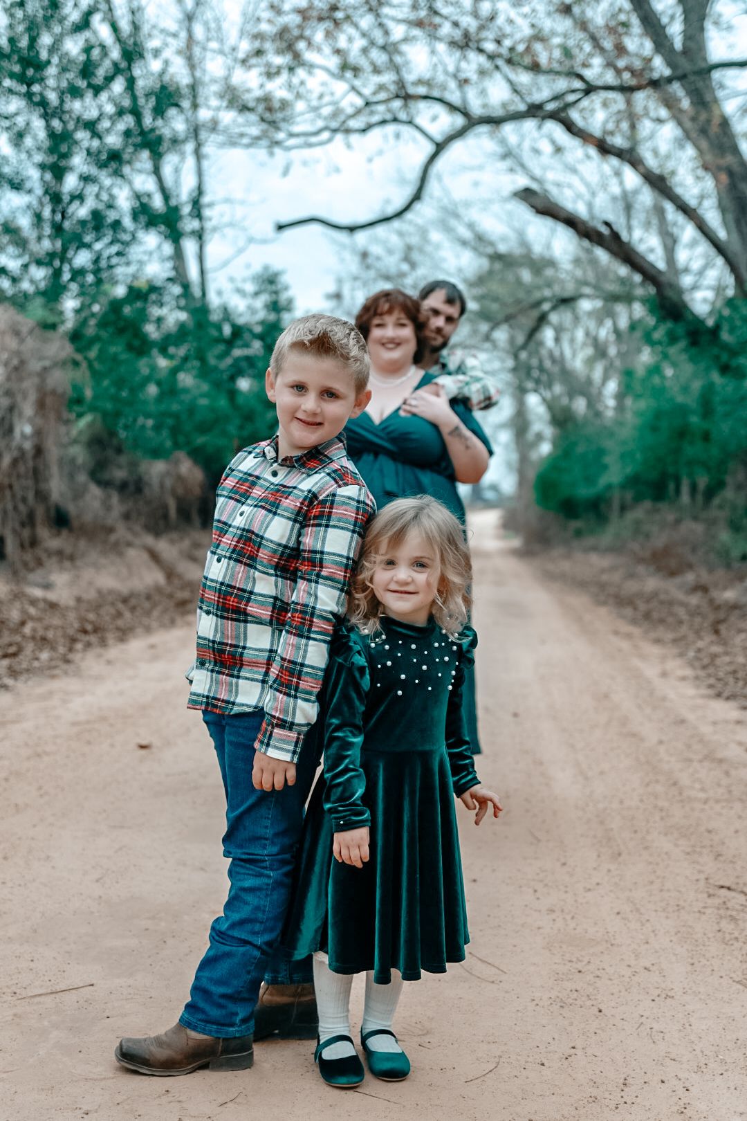A family of four poses on a country dirt road, with two children in front wearing plaid and green velvet and parents behind.