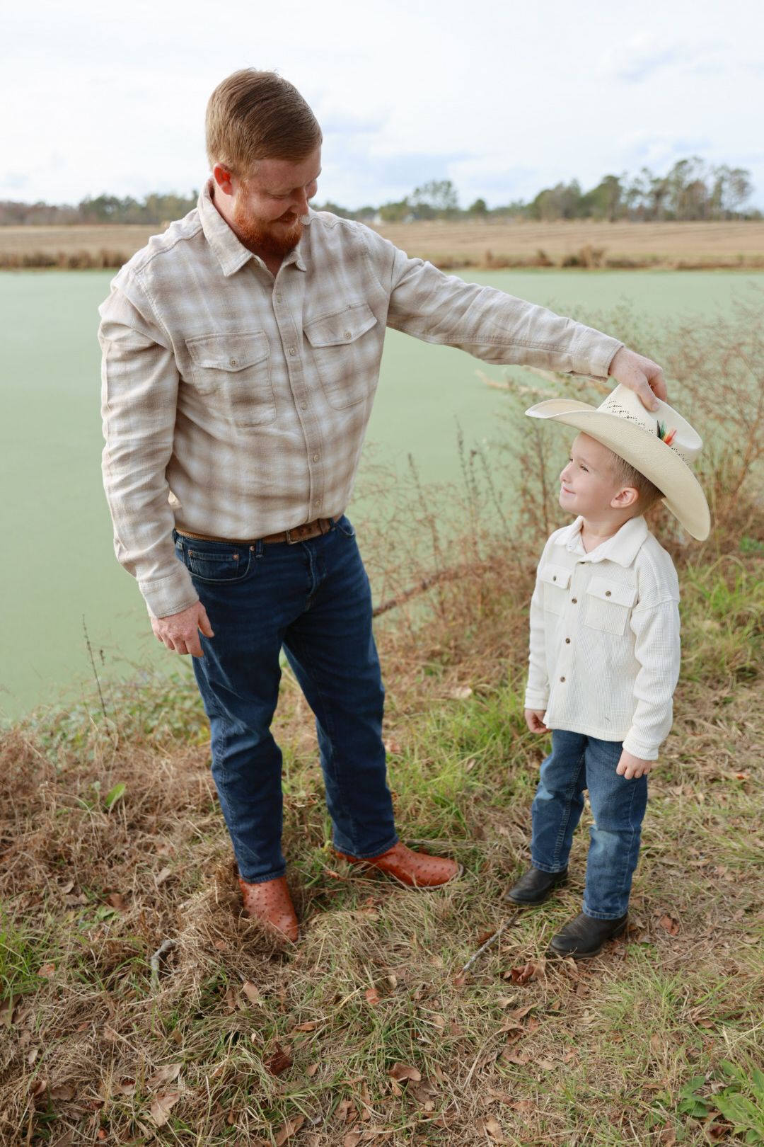 Smiling father in a plaid shirt tips a cowboy hat on his young son beside a green pond in a rustic field.
