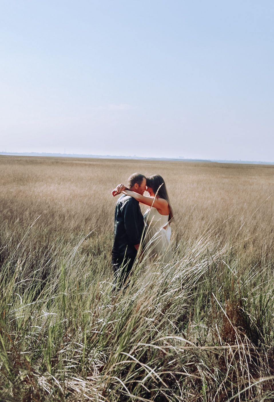 Couple share an embrace in the middle of a vast marsh field of tall grass beneath a hazy blue sky.