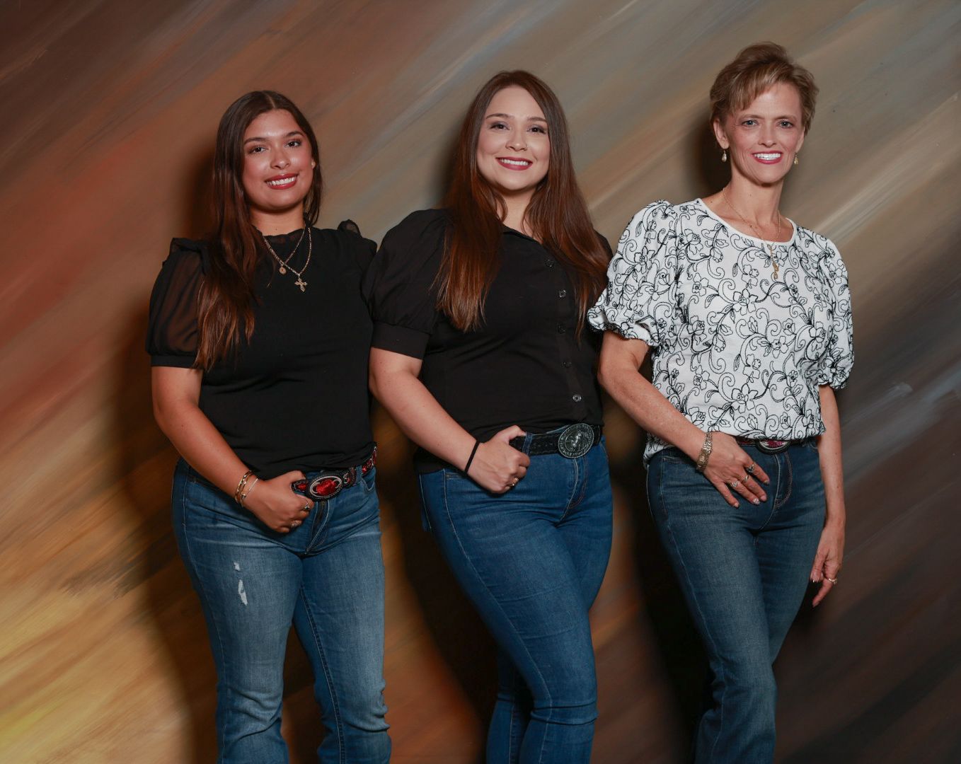 Three smiling women in jeans pose side by side for a studio portrait against a warm, textured backdrop.