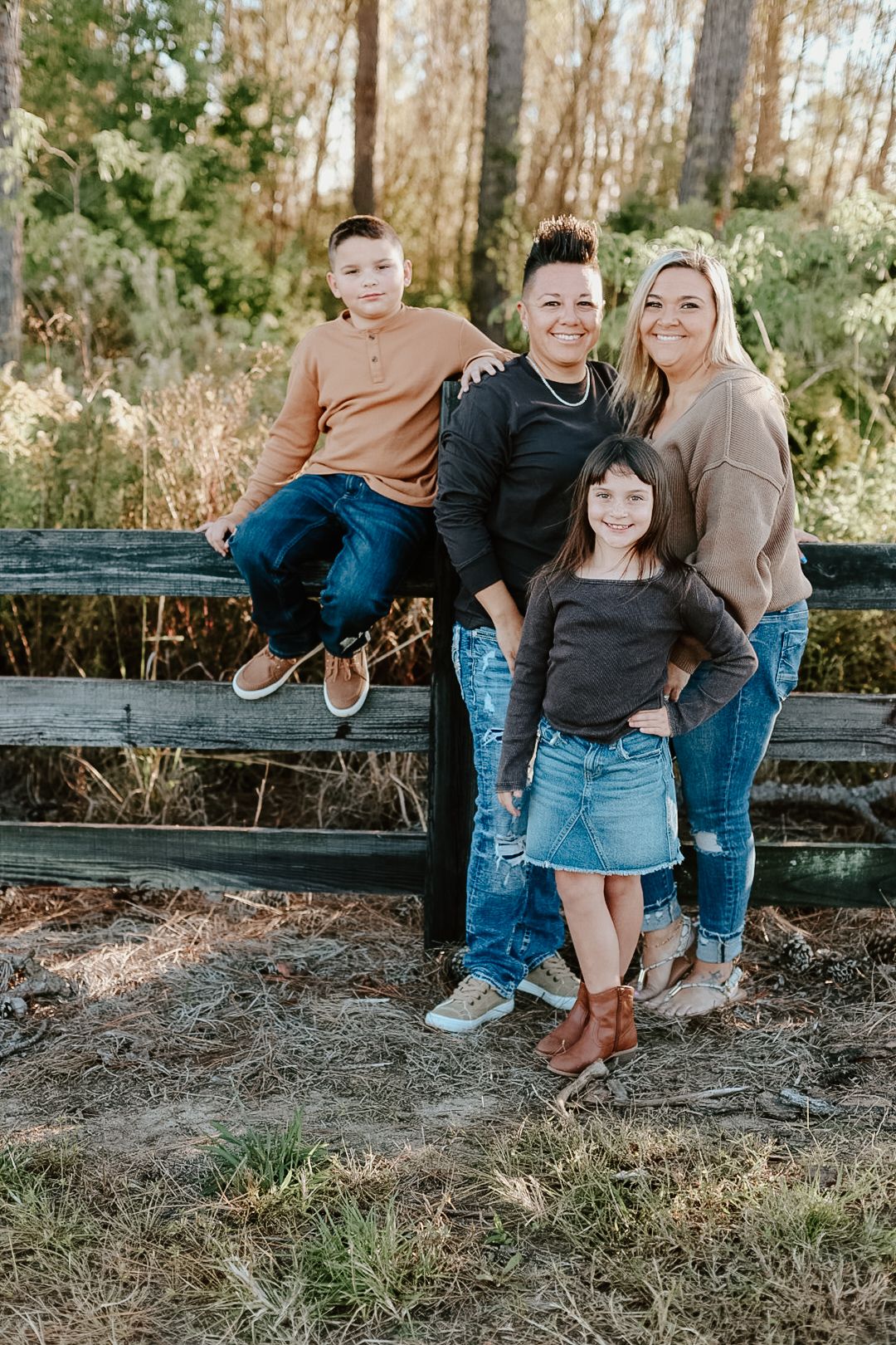 A smiling family of four poses by a wooden fence in a wooded area, with a boy seated on the rail and a girl in front.