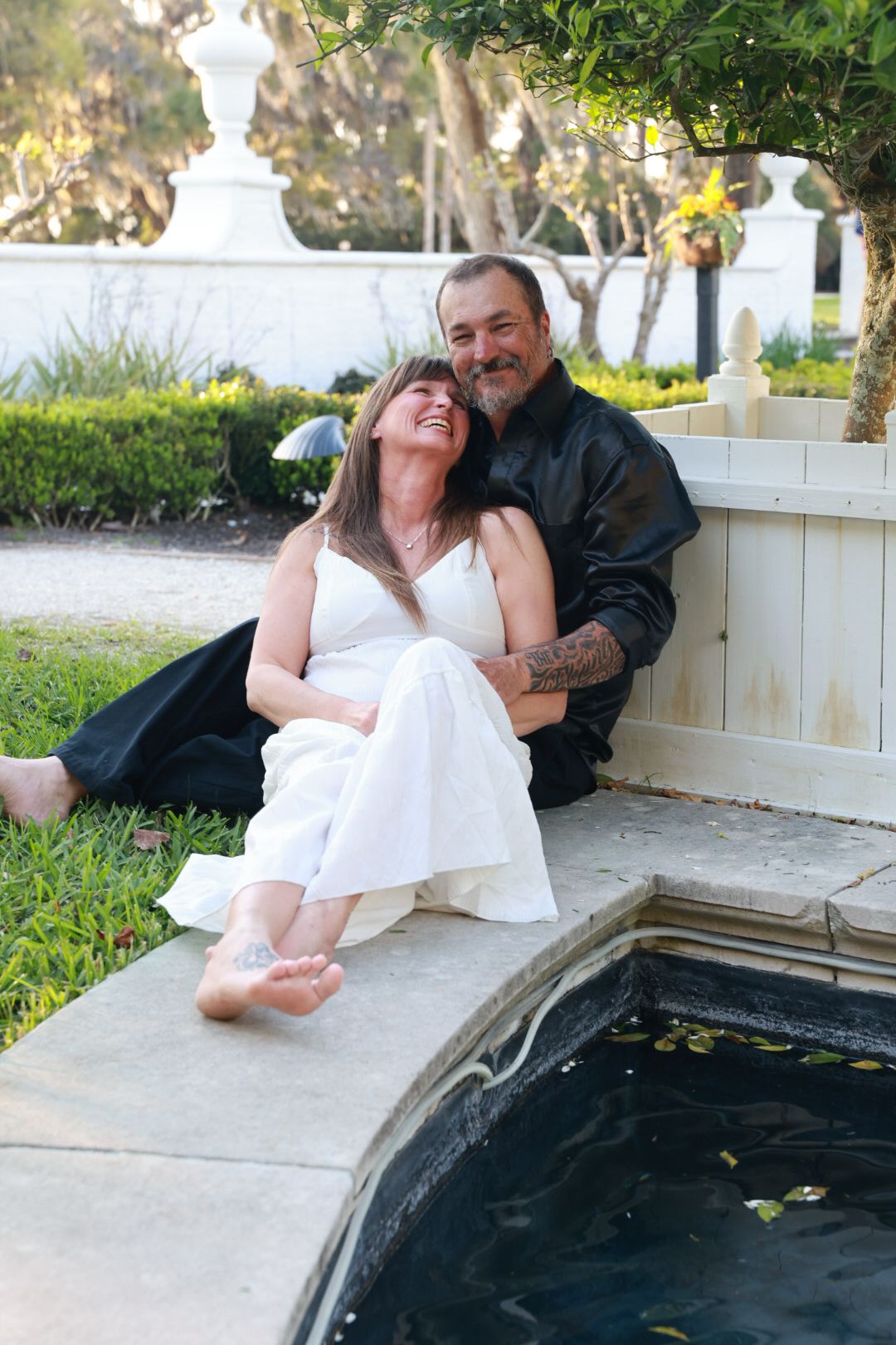 Smiling couple in white dress and black shirt sit laughing together on a stone ledge beside a koi pond.