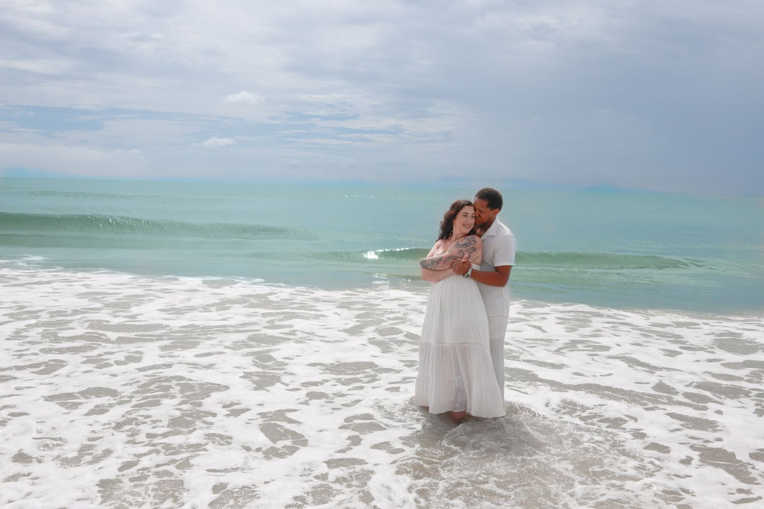 A couple in white embrace while standing in shallow ocean surf under a cloudy sky on the beach.