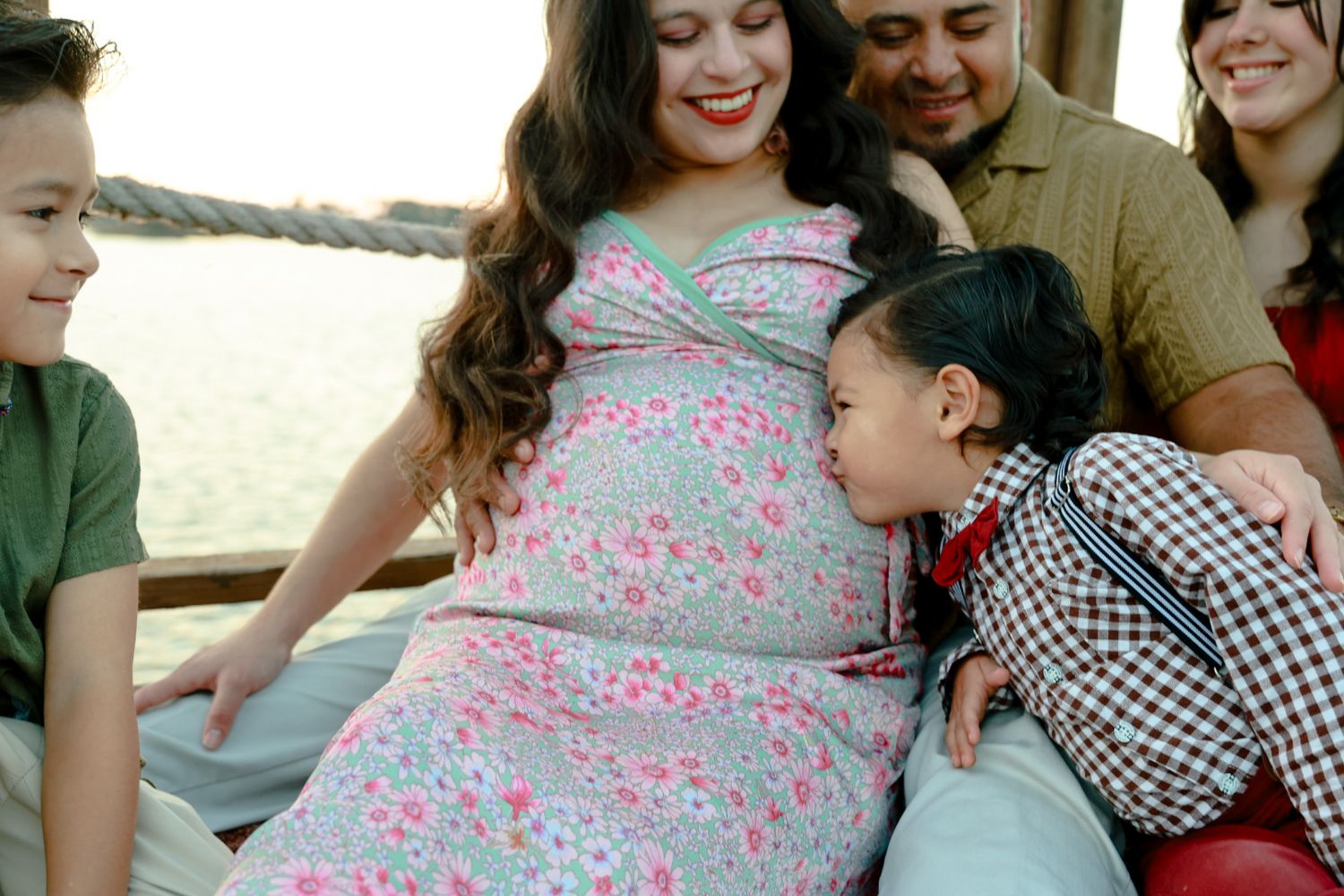 Smiling pregnant woman in a floral dress sits with her husband and children as a little girl kisses her baby bump by the water.