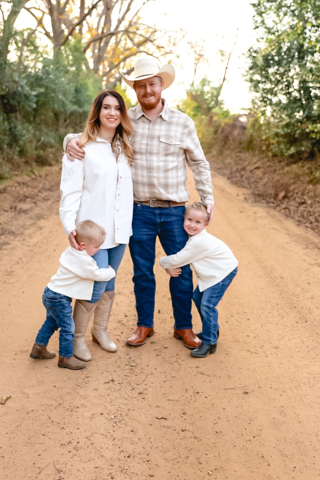 A smiling family of four in western clothing stands on a dirt road, with two young boys hugging their cowboy-hat-wearing parents.