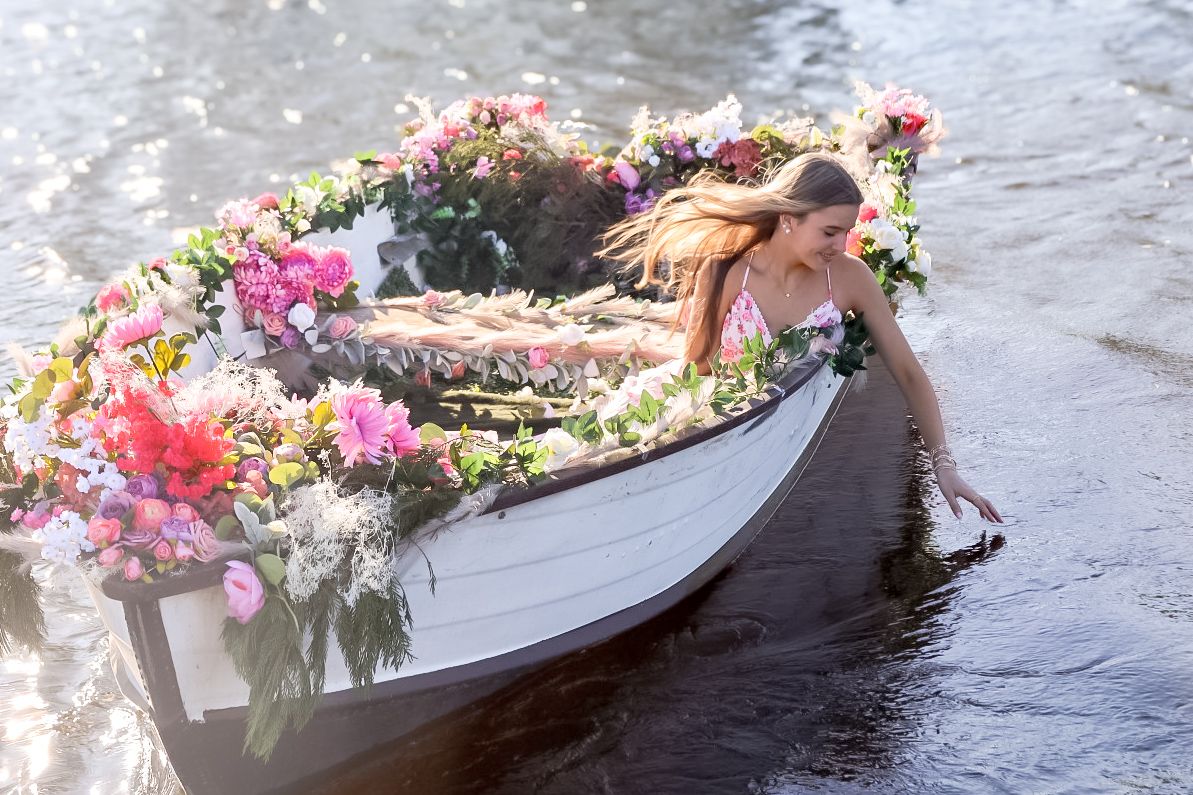 Woman with flowing hair reaching toward the water from a white rowboat decorated with colorful pink and red flowers.