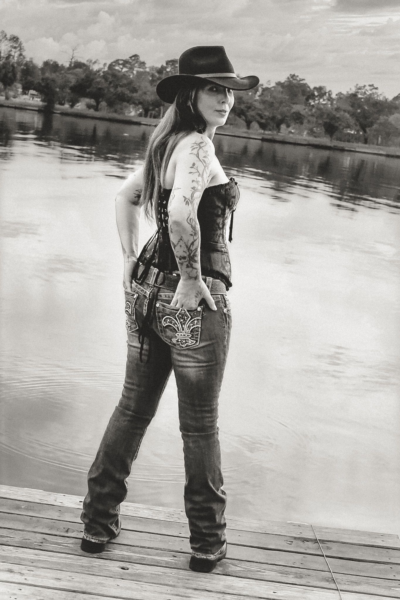 Black and white portrait of a tattooed woman in a cowboy hat, corset top and jeans standing on a wooden dock beside a calm lake.