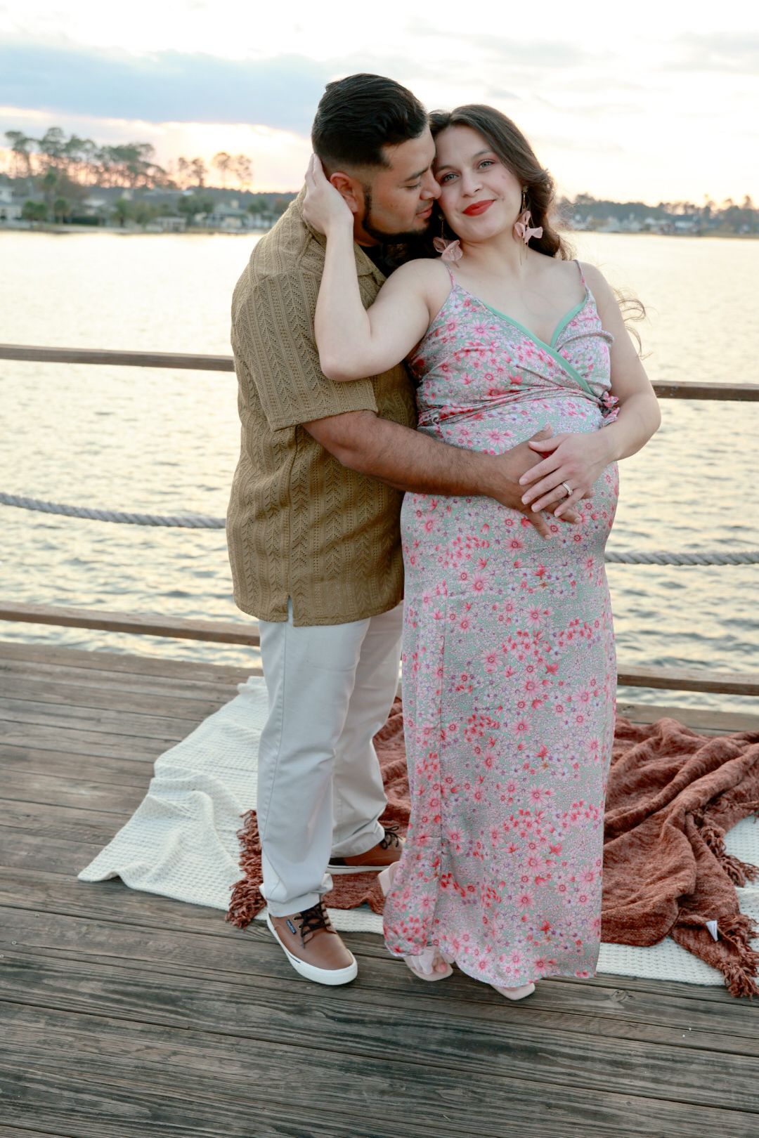 Expectant couple embraces on a wooden lakeside dock at sunset, the man kissing the pregnant woman's cheek as she wears a pink floral dress.