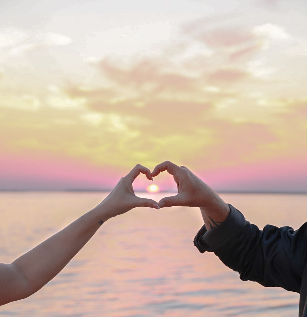 Two hands form a heart shape around the setting sun over the water, with pink and golden clouds in the sky.