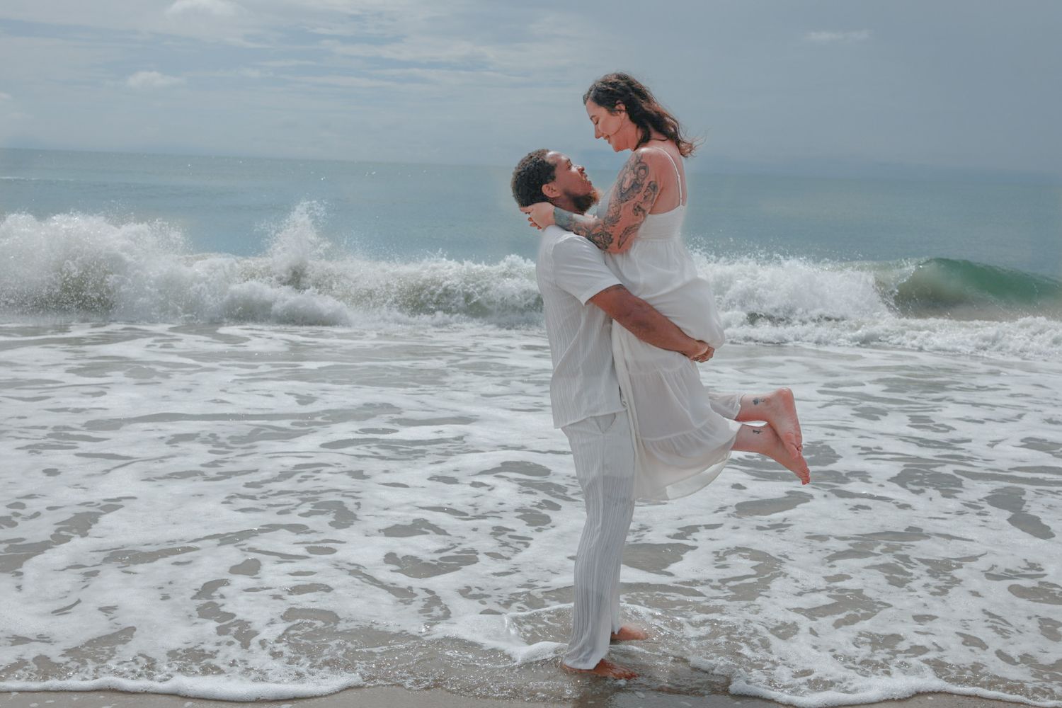 A man in white lifts a barefoot woman in a white dress in his arms as ocean waves crash on the beach behind them.