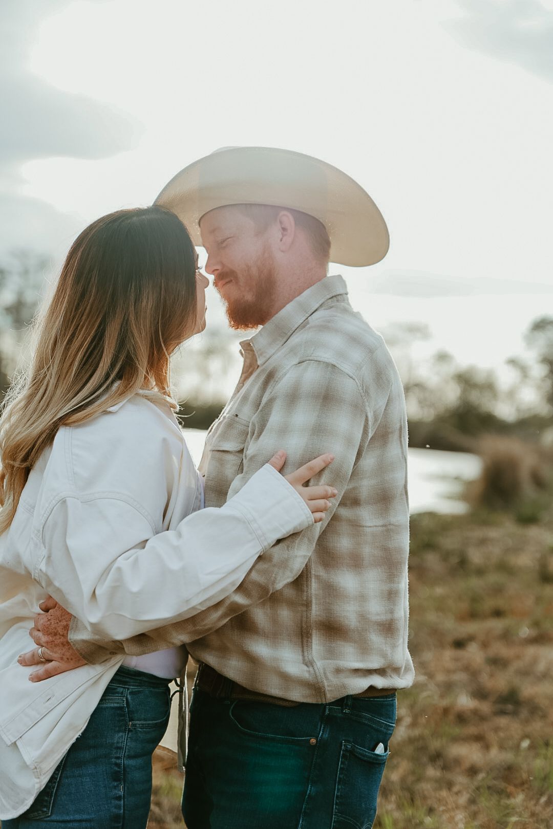 A couple in cowboy hats and casual western wear embrace and touch noses in a soft-lit field near a pond.
