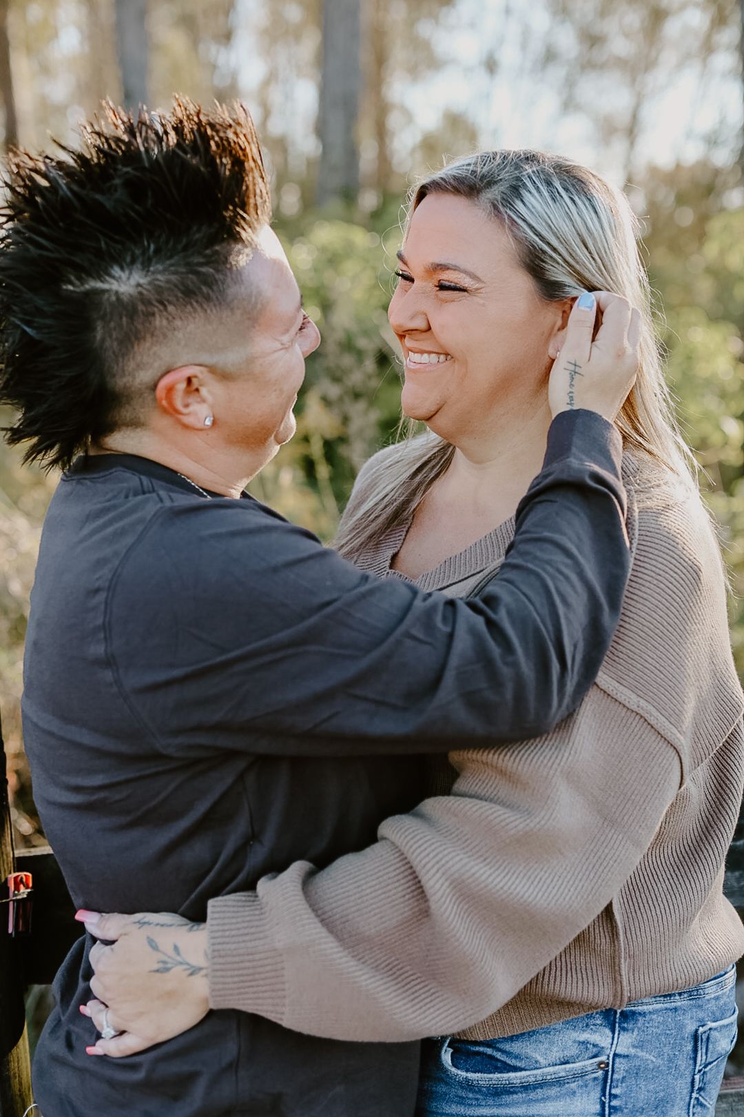 Couple with a black mohawk and long blonde hair smile joyfully as they embrace outdoors in cozy sweaters.