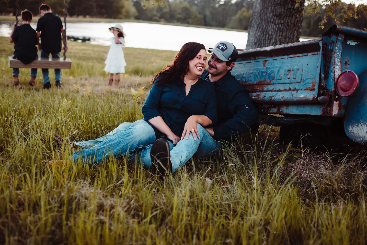 Couple sitting close in tall grass beside a vintage blue truck, with three children playing by the lake behind them.