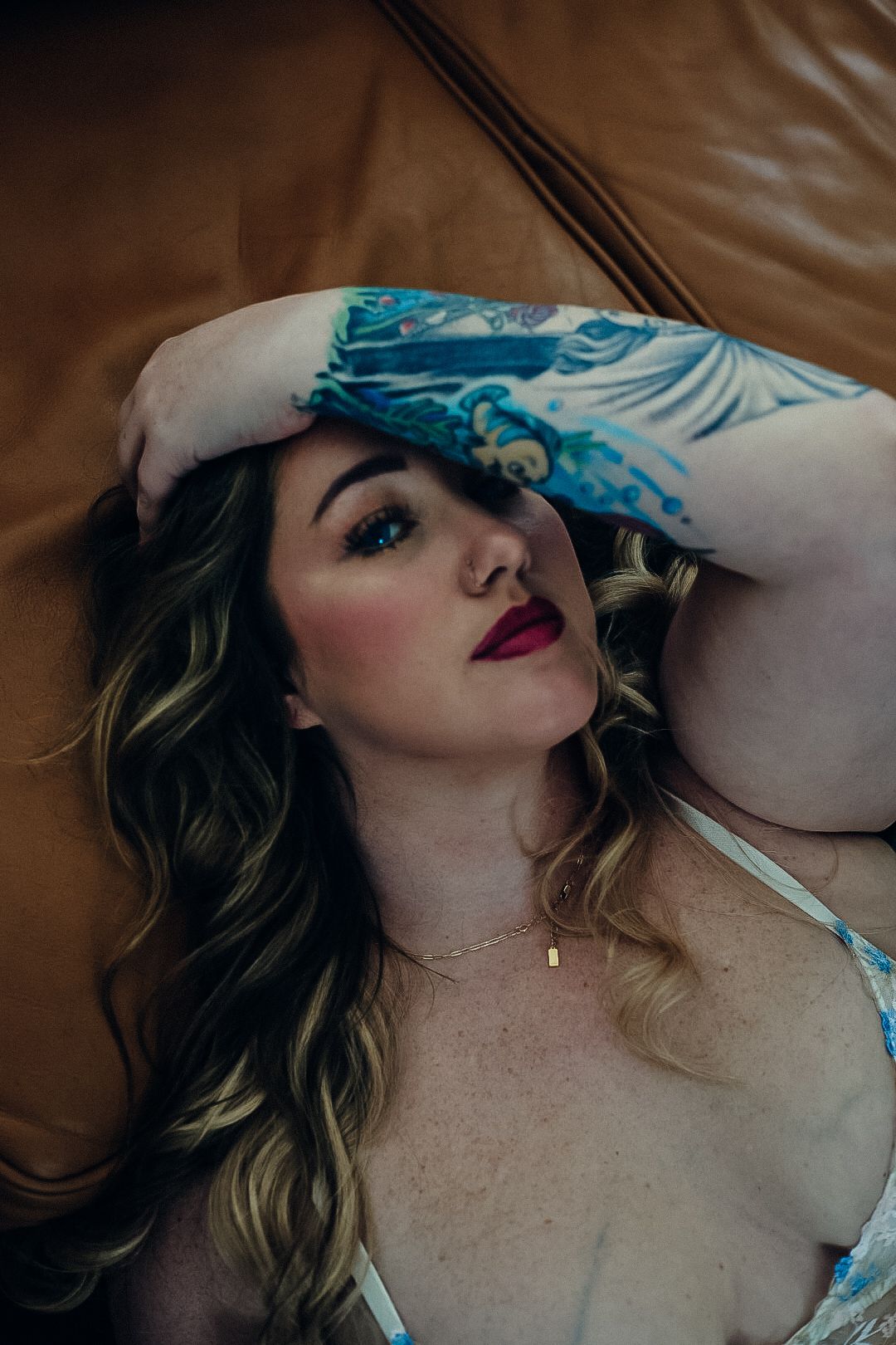 Tattooed woman with wavy blonde hair and red lipstick rests her arm over her head while lying on a tan leather couch.