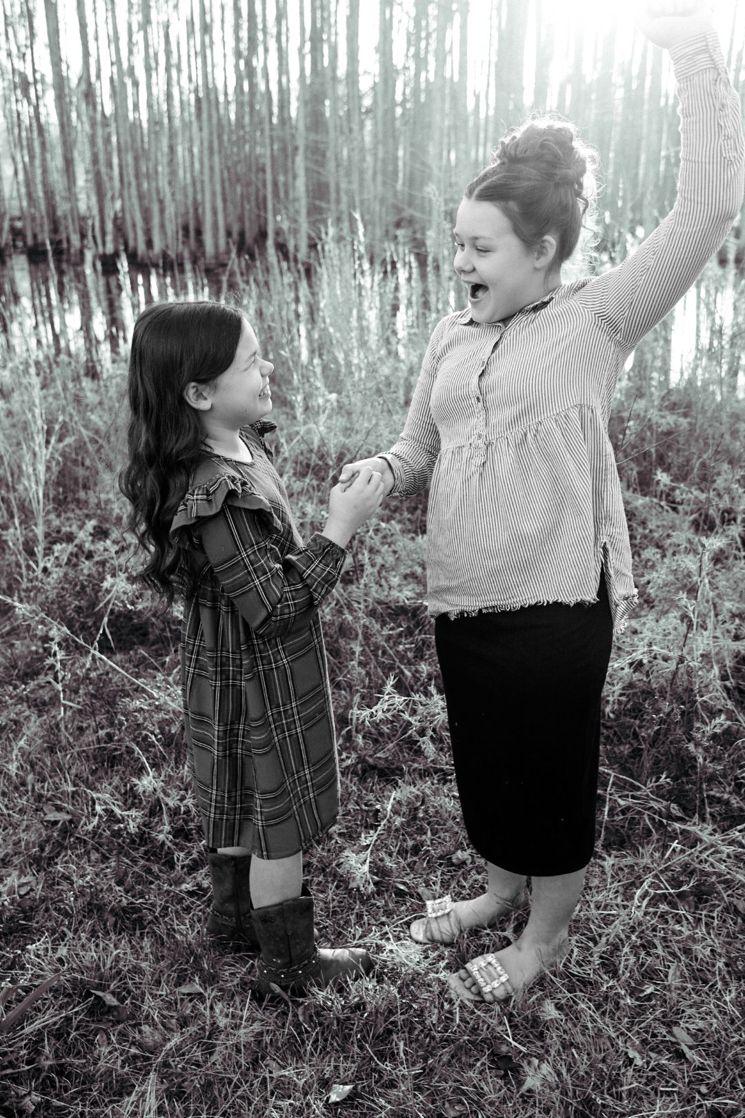 Black and white photo of two girls holding hands and laughing in a grassy field by a woodland pond.
