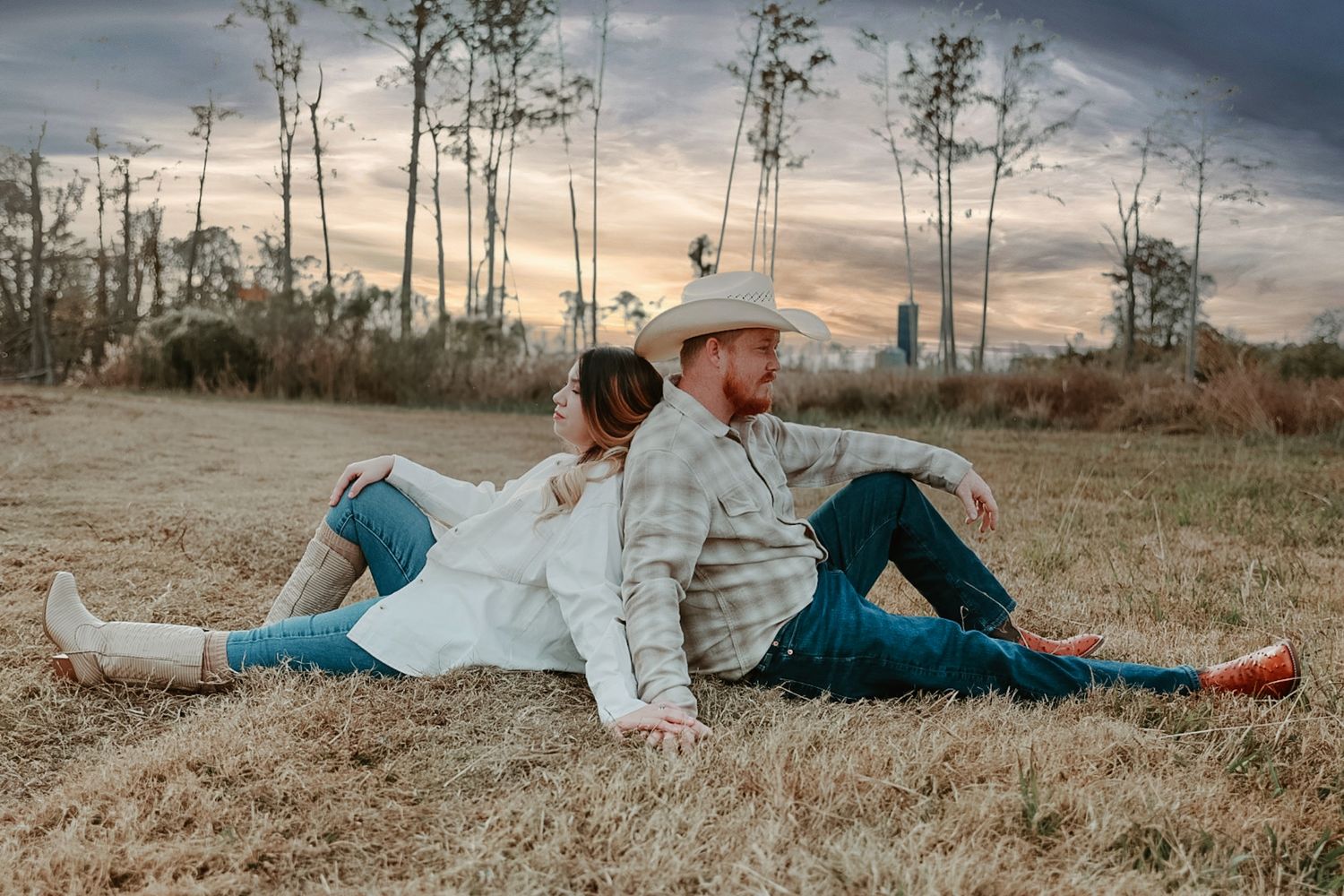 A couple in cowboy hats and boots sit back to back on dry grass at sunset in a rural field with tall pine trees.