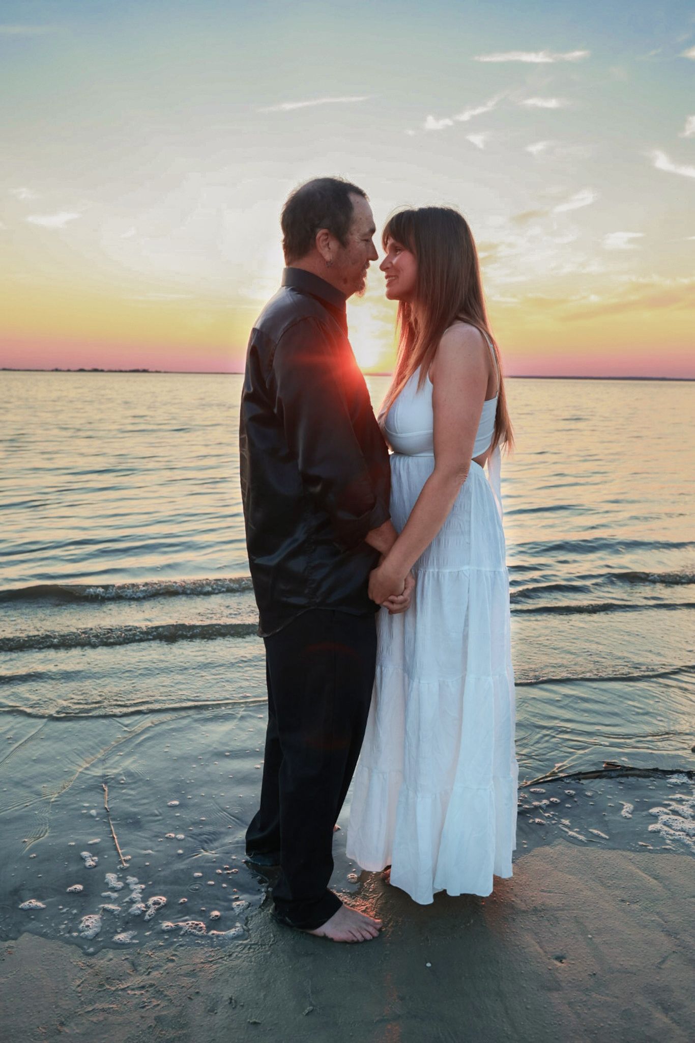 A couple in a black shirt and white dress stand barefoot on the beach, facing each other at sunset beside calm water.