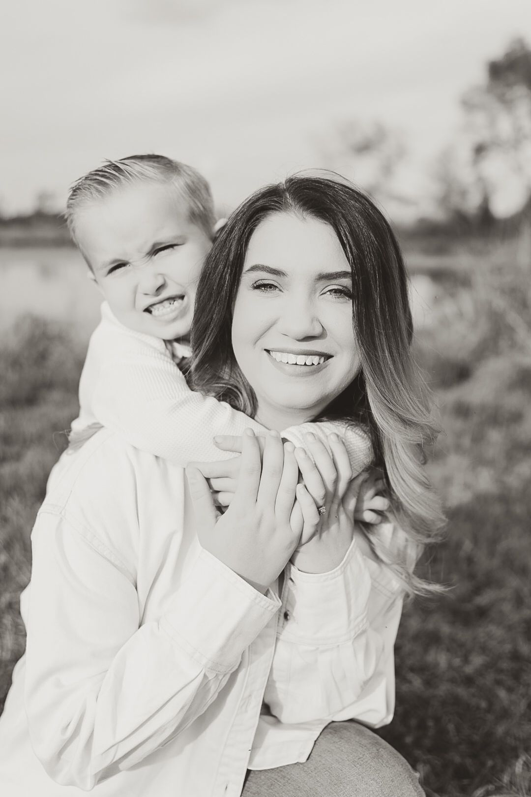 Black and white portrait of a smiling woman in a white shirt with a young boy hugging her from behind outdoors.